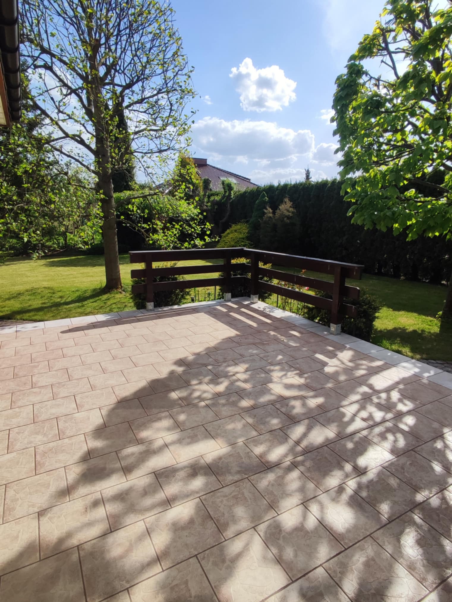 Jasna, wybrukowana kostką tarasowa przestrzeń z drewnianą balustradą, otoczona zielenią ogrodu w słoneczny dzień. Cienie drzew na kostce.