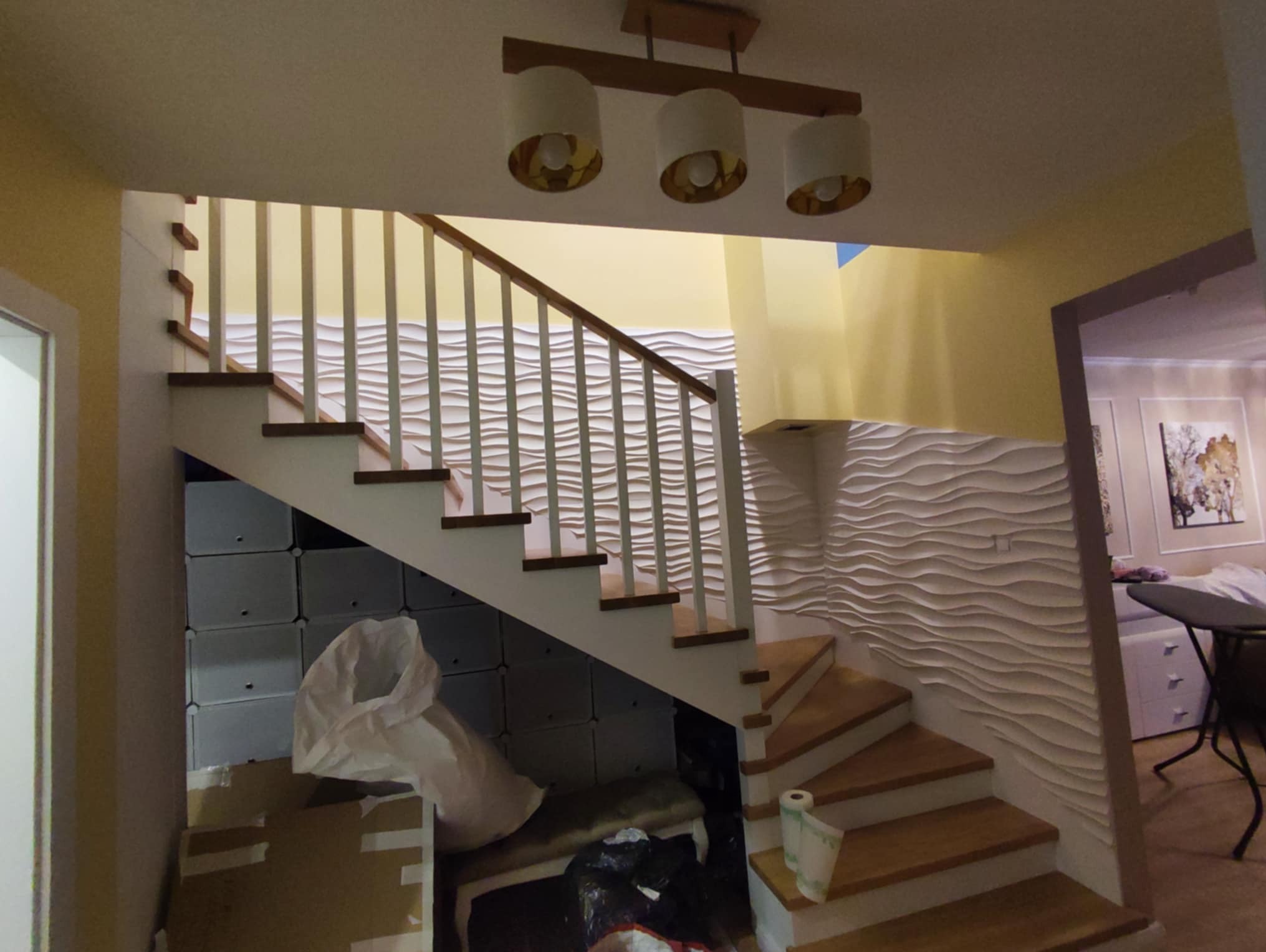 Schody drewniane z białymi balustradami i dekoracyjną ścianą 3D w jasnym wnętrzu. Widoczne oświetlenie sufitowe i elementy wyposażenia.