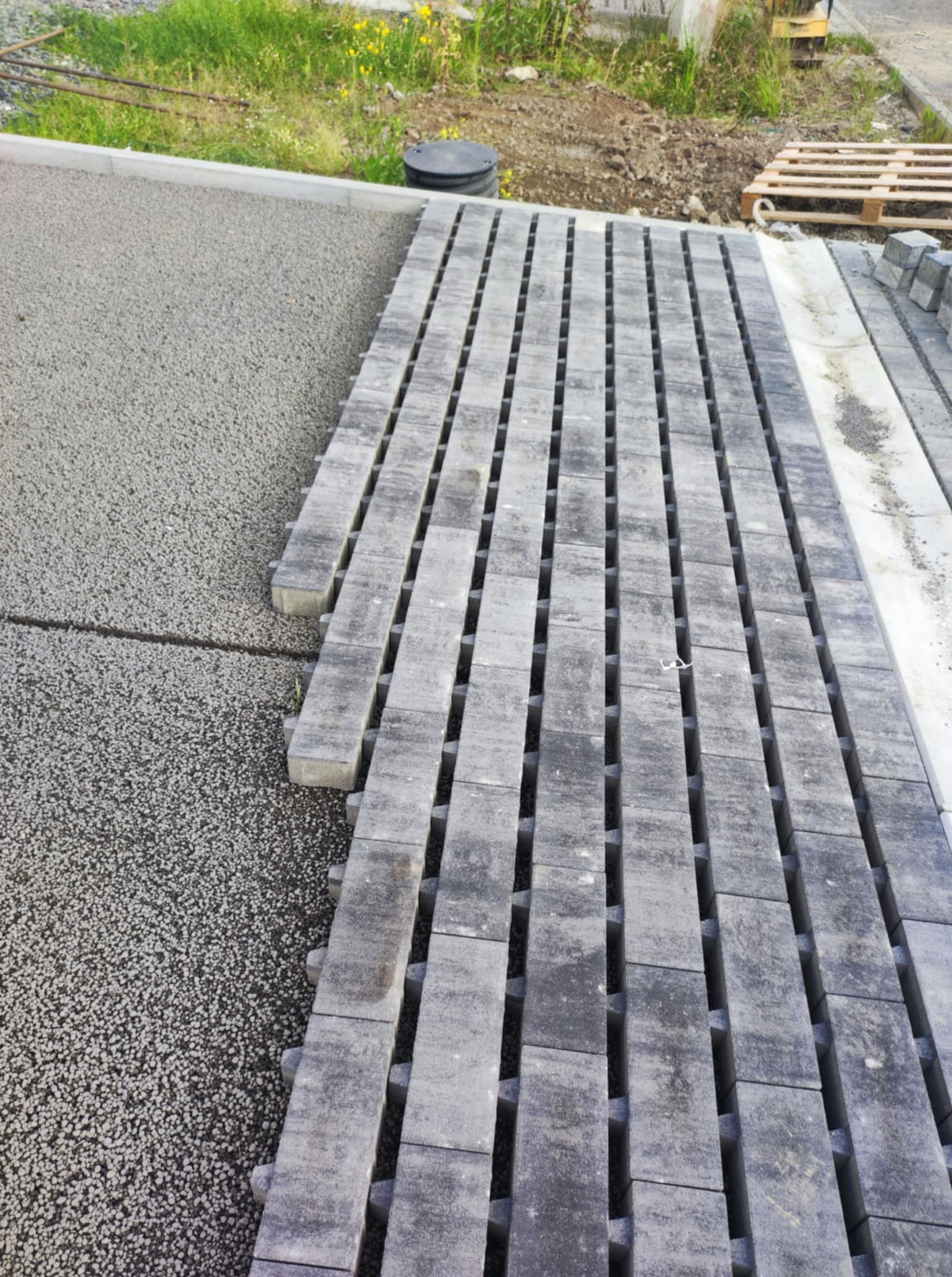 Układanie szarej kostki brukowej na podsypce. Widoczne rzędy kostki z charakterystycznymi przerwami, przygotowane do dalszego układania. Paleta z kostką w tle.