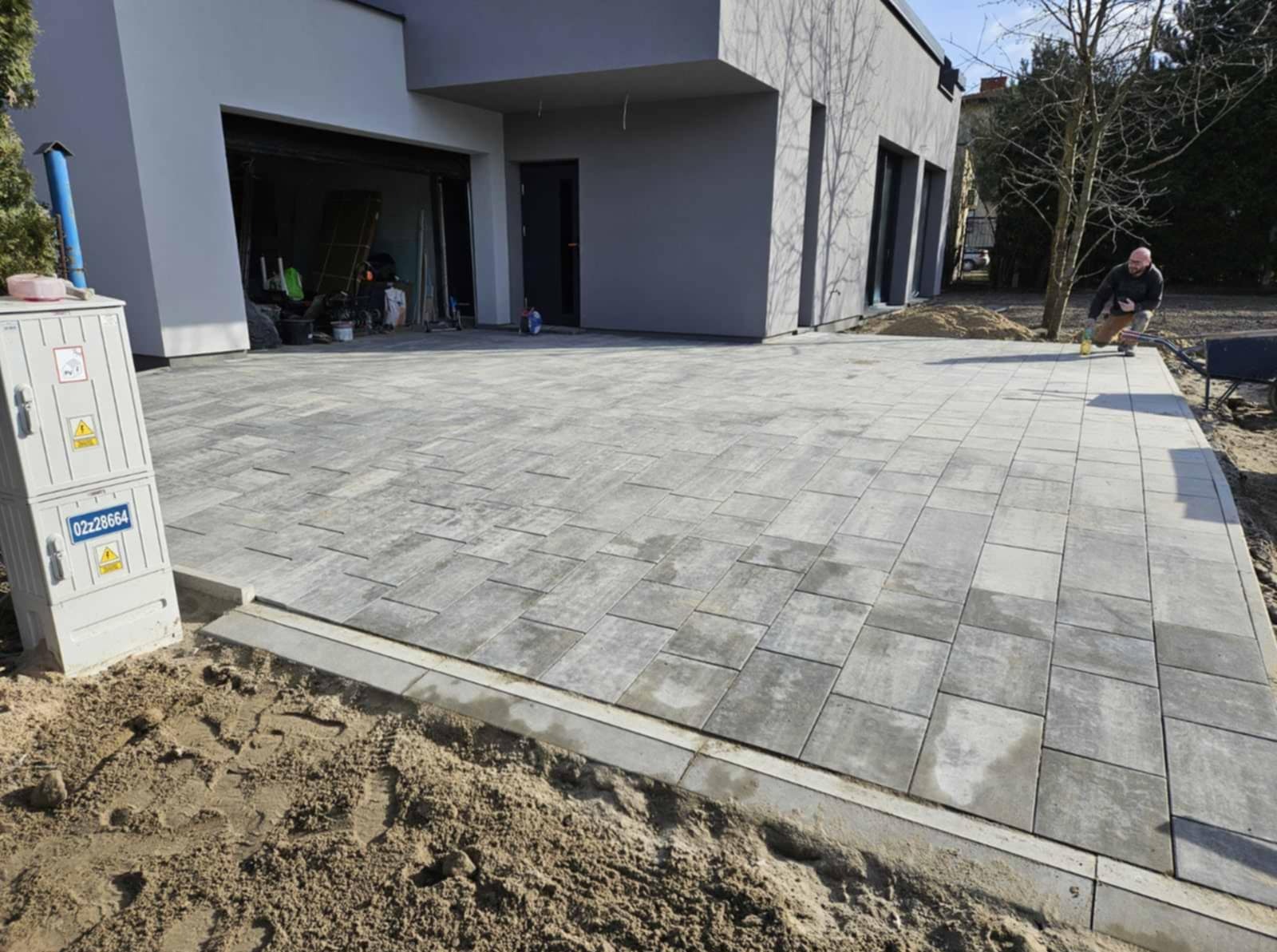 Ułożona szara kostka brukowa na podjeździe nowoczesnego domu. Widoczny fragment elewacji, skrzynka elektryczna i pracownik zamiatający powierzchnię.