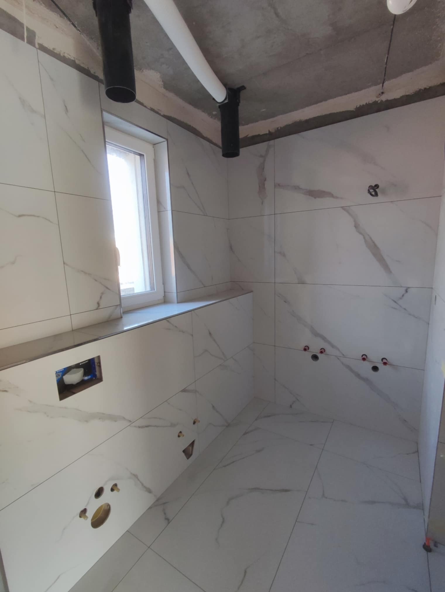 Wykończona łazienka z białymi płytkami imitującymi marmur, widoczne rury i instalacje podtynkowe. Okno z parapetem. Surowy betonowy sufit.