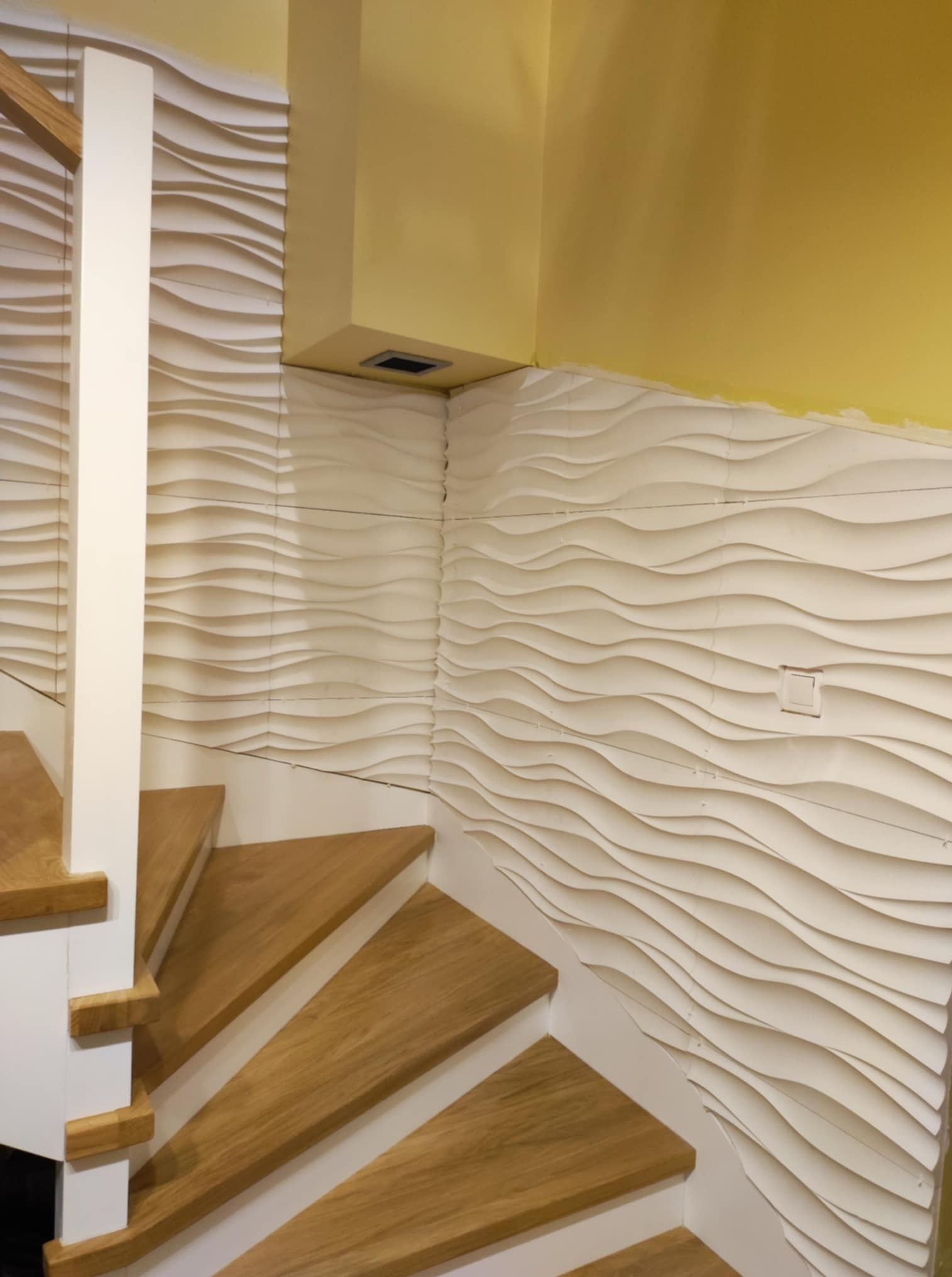 Schody z drewnianymi stopniami i dekoracyjną, białą ścianą 3D w Piasecznie. Żółta ściana w tle. Detal wykończenia wnętrza.