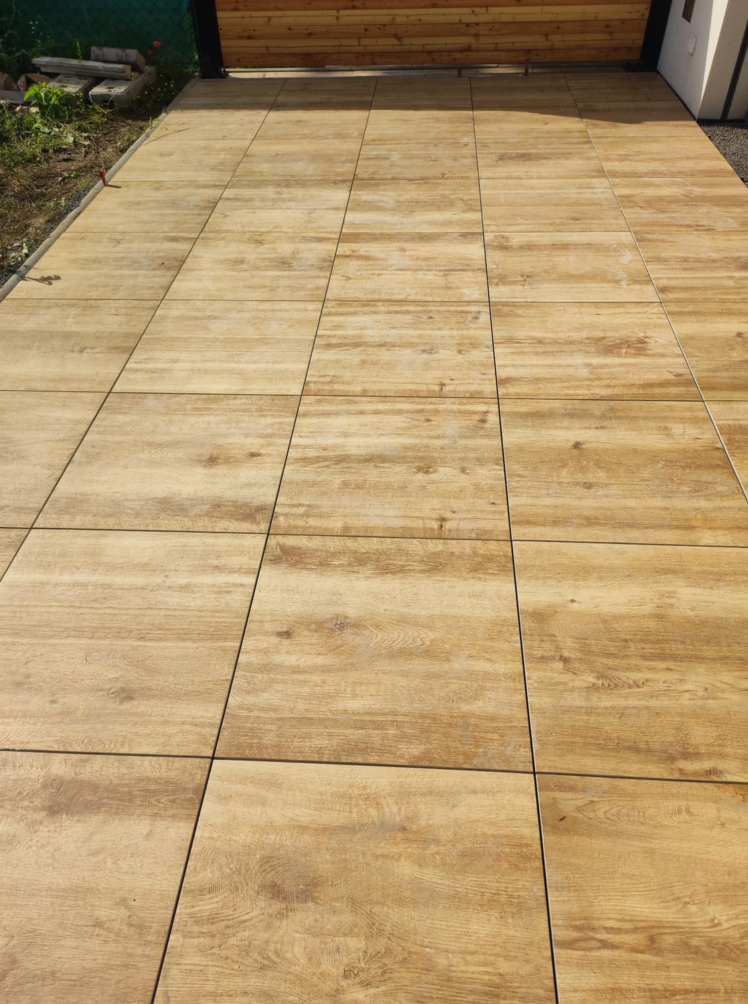 Ułożona, jasnobrązowa, kwadratowa kostka imitująca drewno na podjeździe. Widok z góry, regularny wzór, fuga w kolorze czarnym. Częściowo widoczna zieleń i elementy konstrukcyjne.