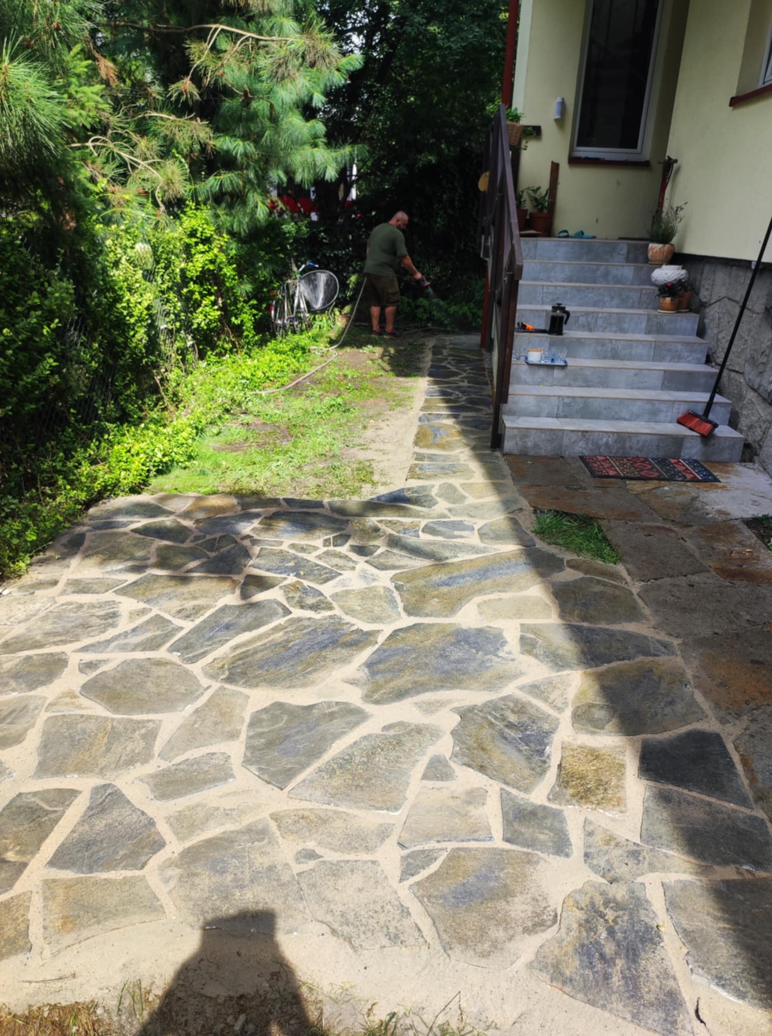 Ścieżka z nieregularnych kamieni prowadząca do schodów domu, z widocznym mężczyzną czyszczącym ją myjką ciśnieniową, otoczona zielenią.