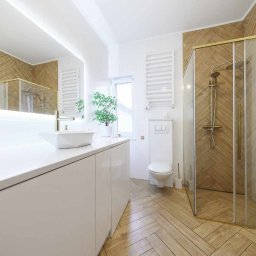 Nowoczesna łazienka z drewnopodobnymi płytkami ułożonymi w jodełkę na podłodze i ścianie prysznica, białą armaturą i złotymi akcentami.