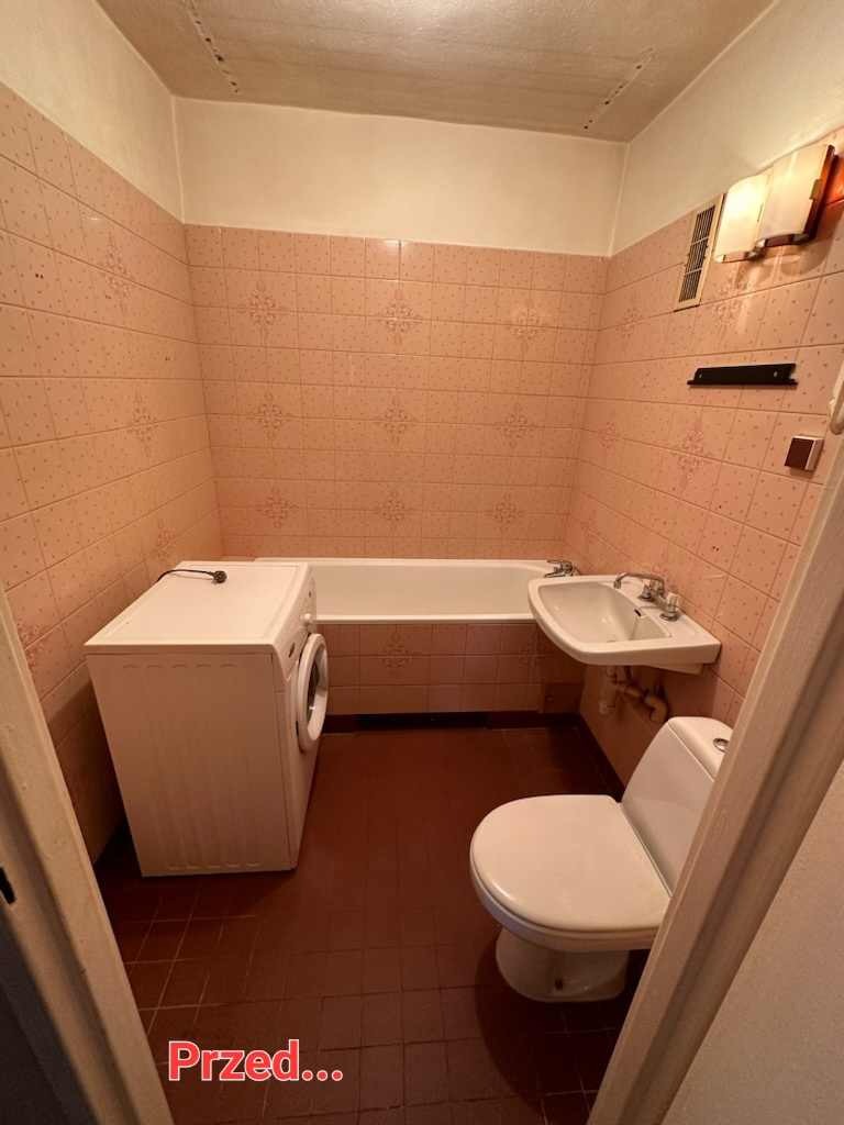 Łazienka w różowych płytkach przed remontem: widok na pralkę, wannę, umywalkę i toaletę. Ciemna podłoga z płytek. Widoczny fragment drzwi.