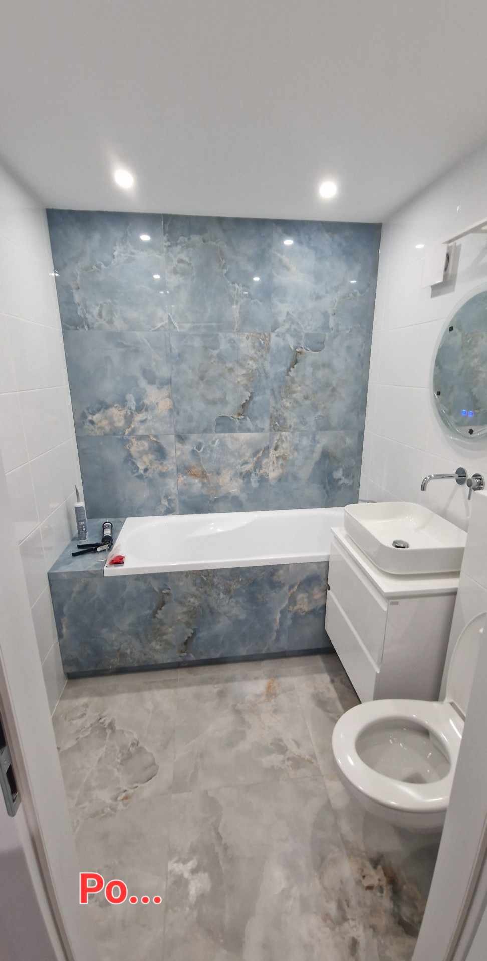 Wykończona łazienka z wanną obudowaną płytkami imitującymi marmur, umywalką nablatową i toaletą. Podłoga wyłożona płytkami w podobnym stylu. Oświetlenie punktowe.