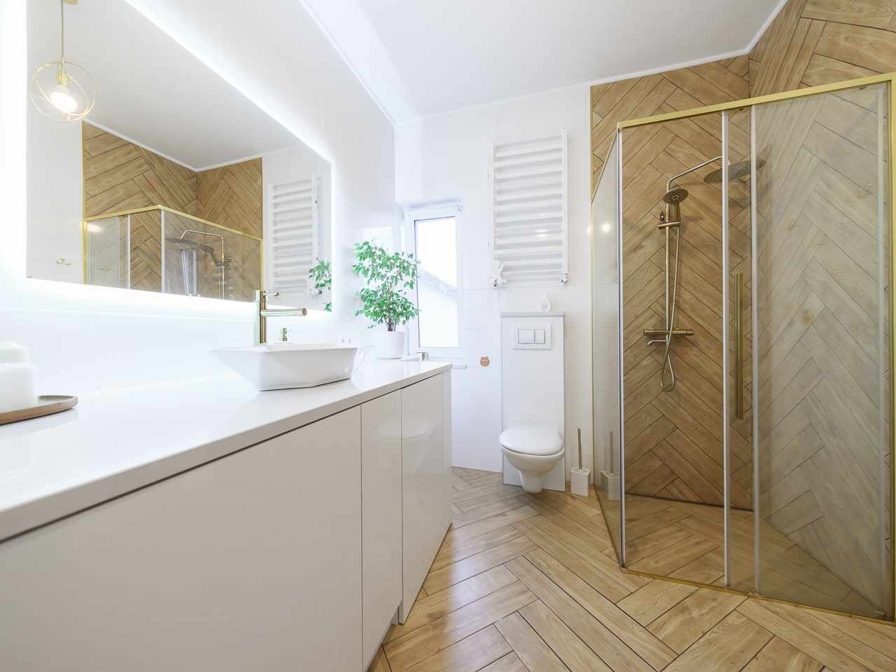 Nowoczesna łazienka z drewnopodobnymi płytkami ułożonymi w jodełkę na podłodze i ścianie prysznica, białą armaturą i złotymi akcentami.