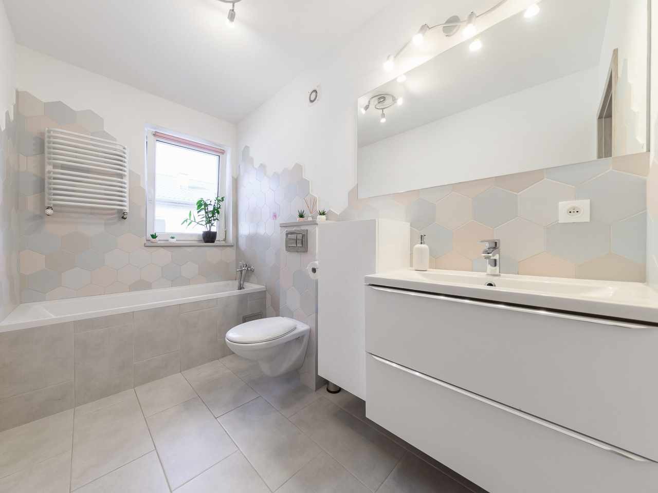 Jasna łazienka z wanną, umywalką i heksagonalnymi płytkami w odcieniach szarości i różu. Minimalistyczny design, nowoczesne oświetlenie i subtelne akcenty roślinne.