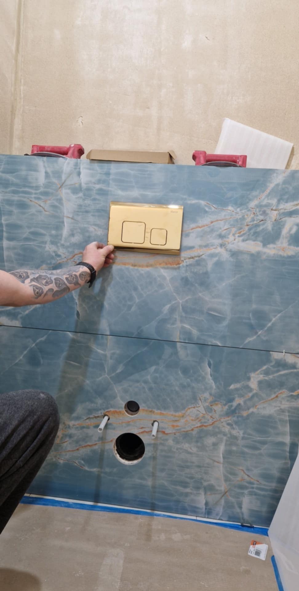 Błękitna ściana z marmurowym wzorem w łazience, z otworem na muszlę klozetową i przyciskiem spłukującym w kolorze złotym, trzymanym przez osobę z tatuażem na ramieniu.