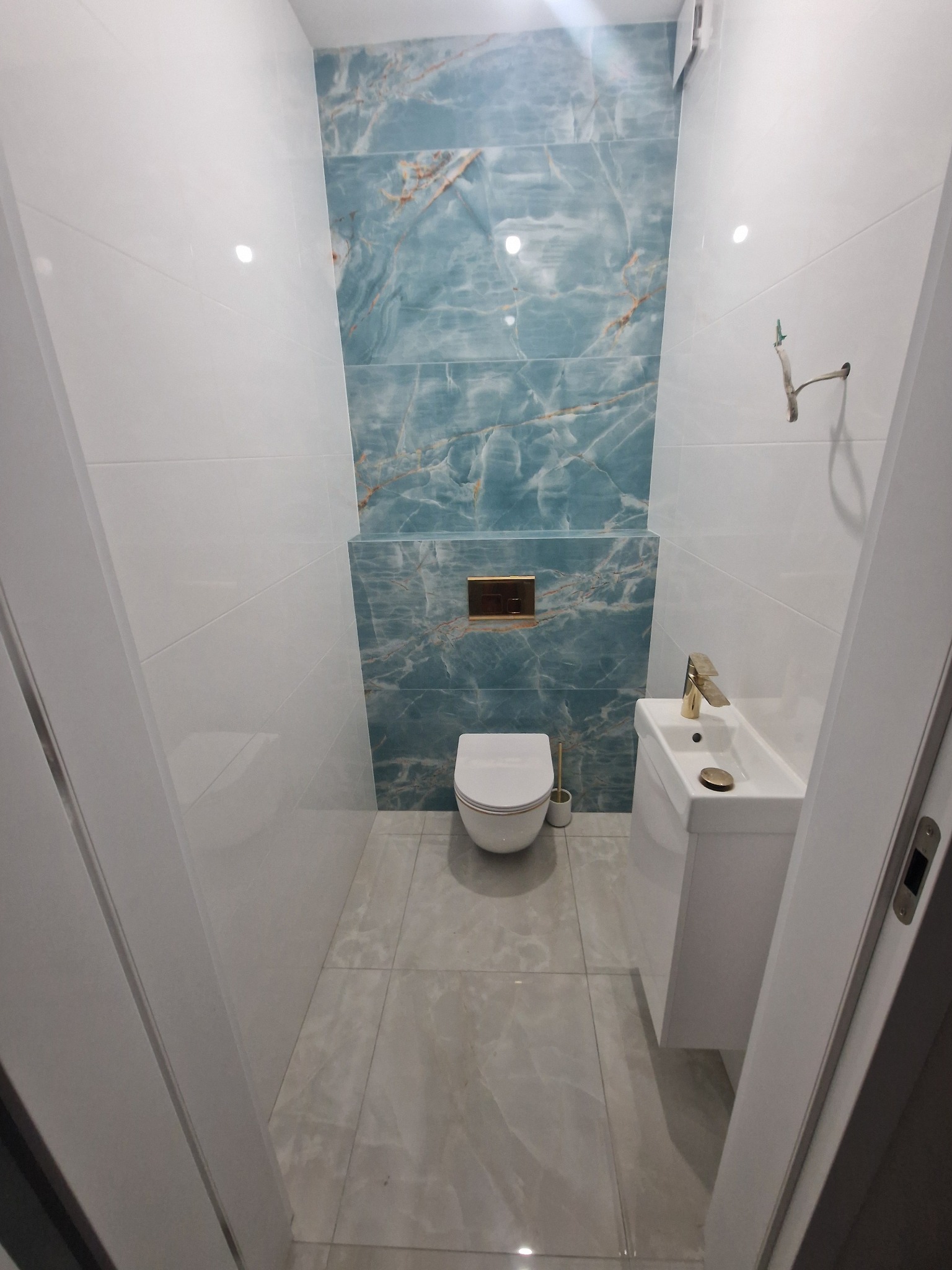 Wykończona toaleta z białą armaturą i umywalką, akcentowana płytkami imitującymi marmur w odcieniach błękitu i złota. Złota armatura i detale dodają elegancji.