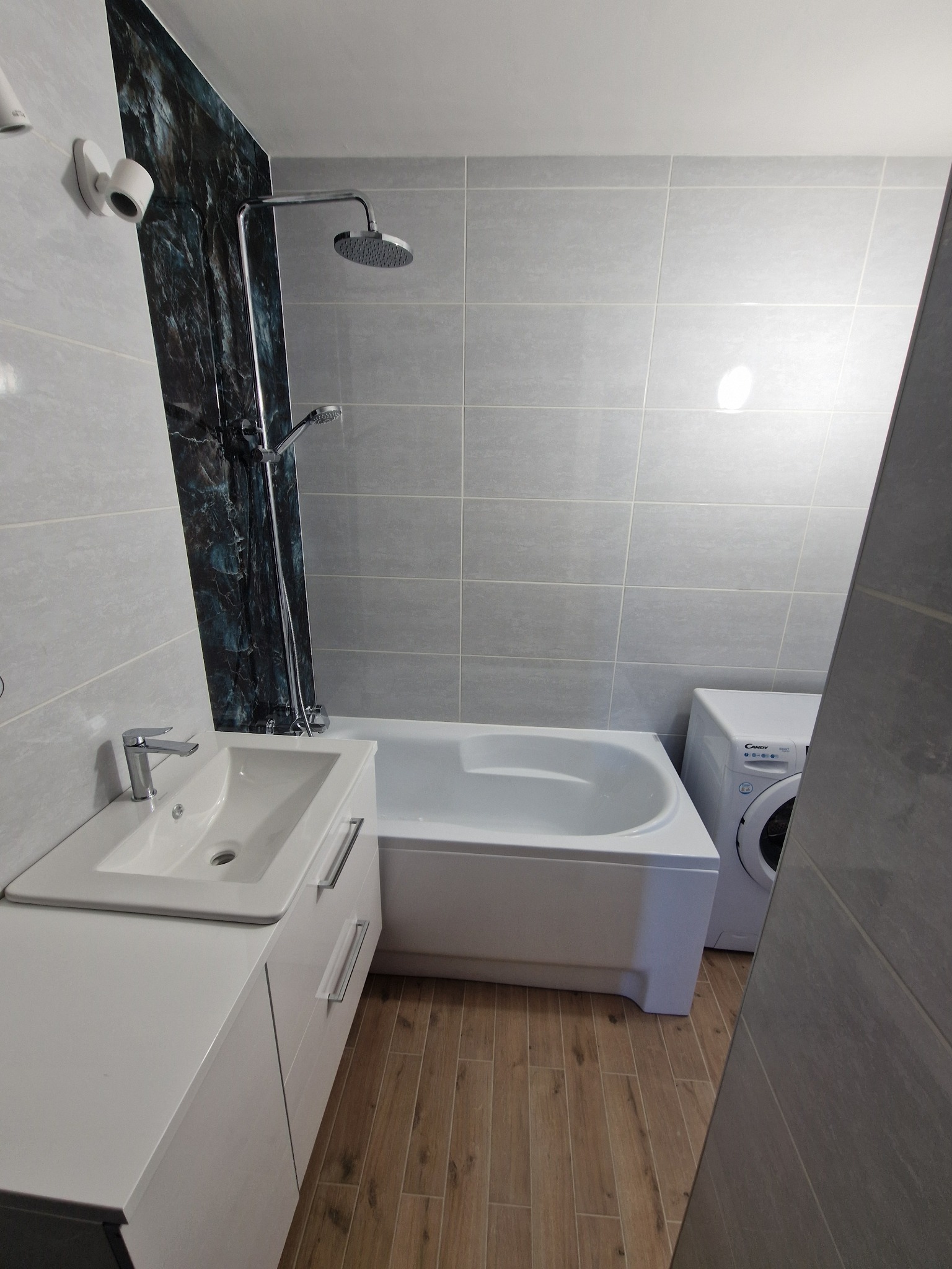 Nowoczesna łazienka po remoncie: szare płytki, umywalka z szafką, wanna i pralka. Akcent stanowi panel dekoracyjny imitujący marmur przy prysznicu.