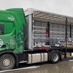 Vervo - Ciężarówka marki Scania firmy Vervo z otwartą naczepą, w której widać zabezpieczony ładunek metalowych elementów. Zielono-biała kolorystyka pojazdu.