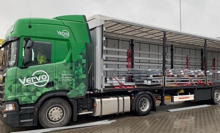 Ciężarówka marki Scania firmy Vervo z otwartą naczepą, w której widać zabezpieczony ładunek metalowych elementów. Zielono-biała kolorystyka pojazdu.