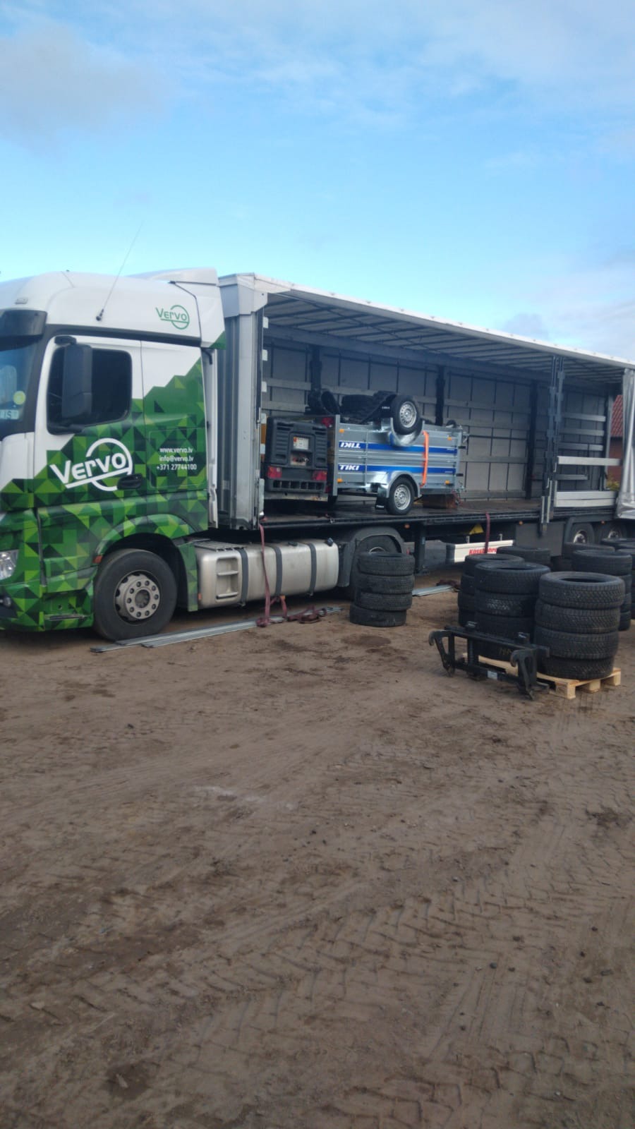 Ciężarówka firmy Vervo z przyczepą i agregatem wewnątrz, obok sterty opon. Widoczne logo firmy i dane kontaktowe na kabinie. Transport ładunków.