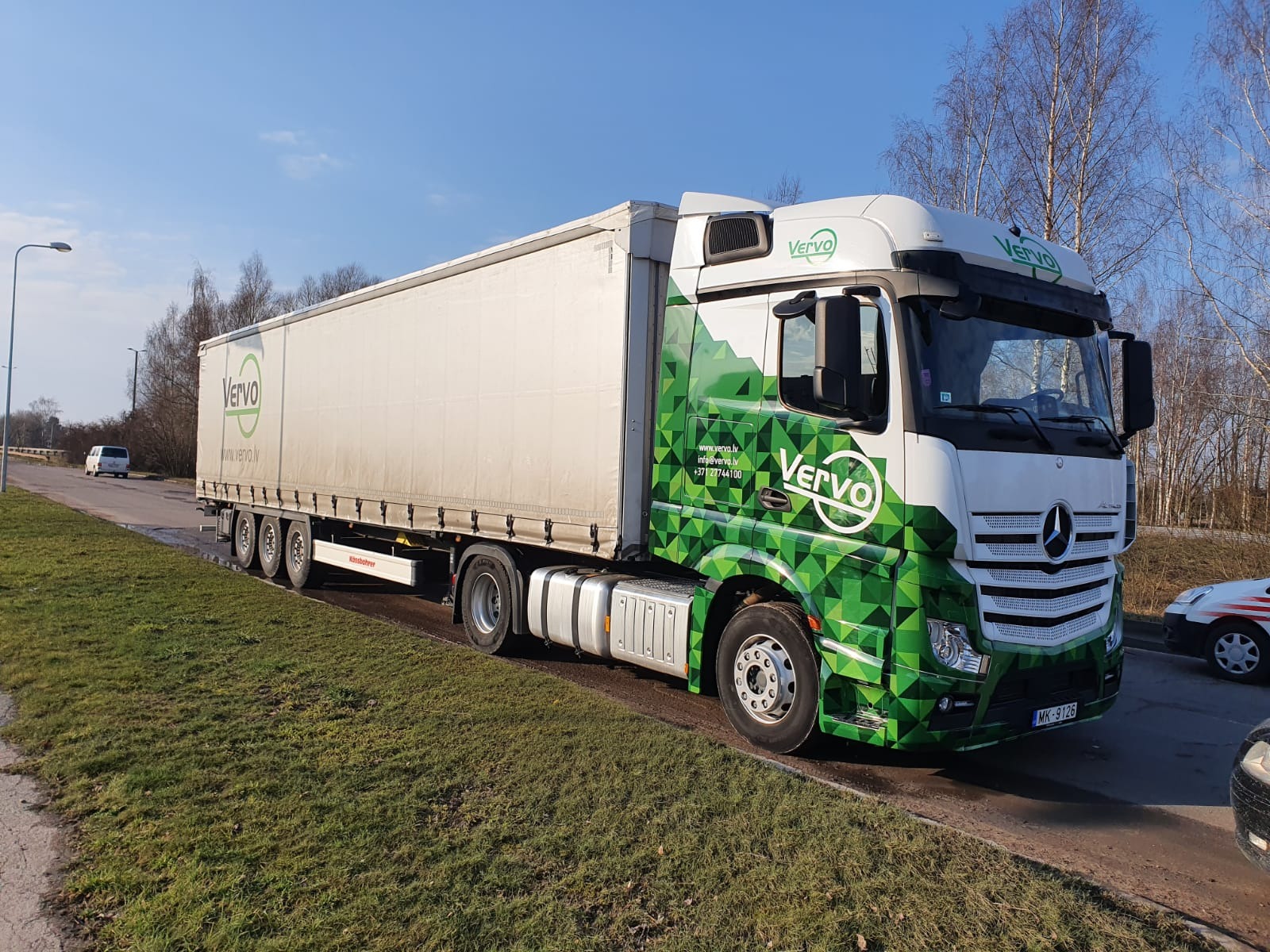 Ciężarówka marki Mercedes-Benz z naczepą, oklejona w zielono-białe wzory geometryczne i logo firmy Vervo, zaparkowana na poboczu drogi w słoneczny dzień.