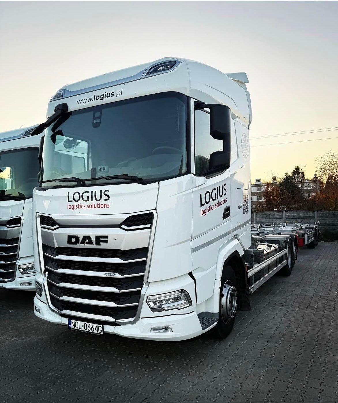 Biały ciągnik siodłowy DAF z naczepą kontenerową i logo Logius na parkingu z kostki brukowej, widok z przodu, tablica rejestracyjna widoczna.