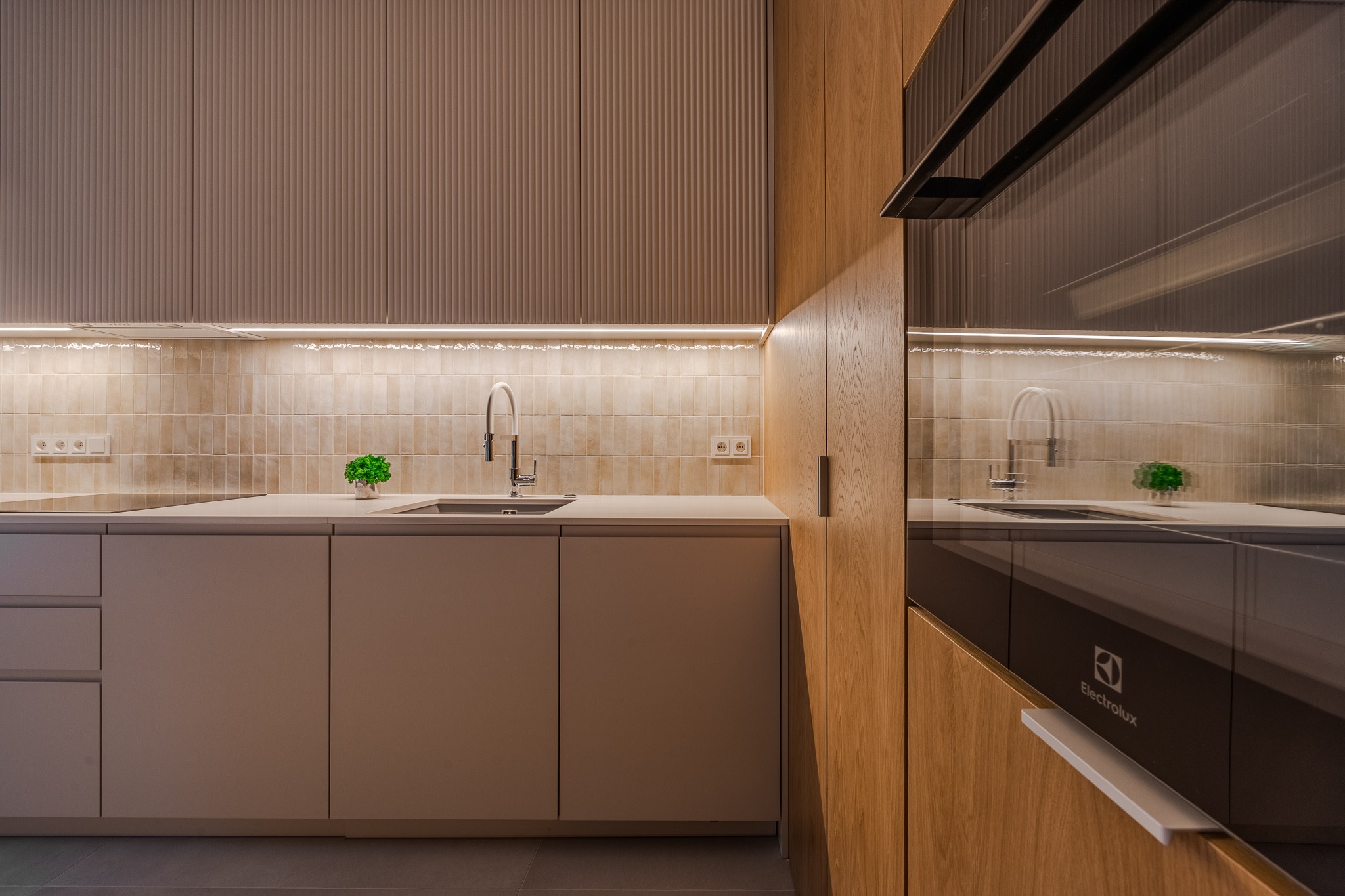 Nowoczesna kuchnia z beżowymi frontami szafek z ryflowanym wzorem, oświetleniem LED podszafkowym i sprzętem Electrolux. Minimalistyczny design.