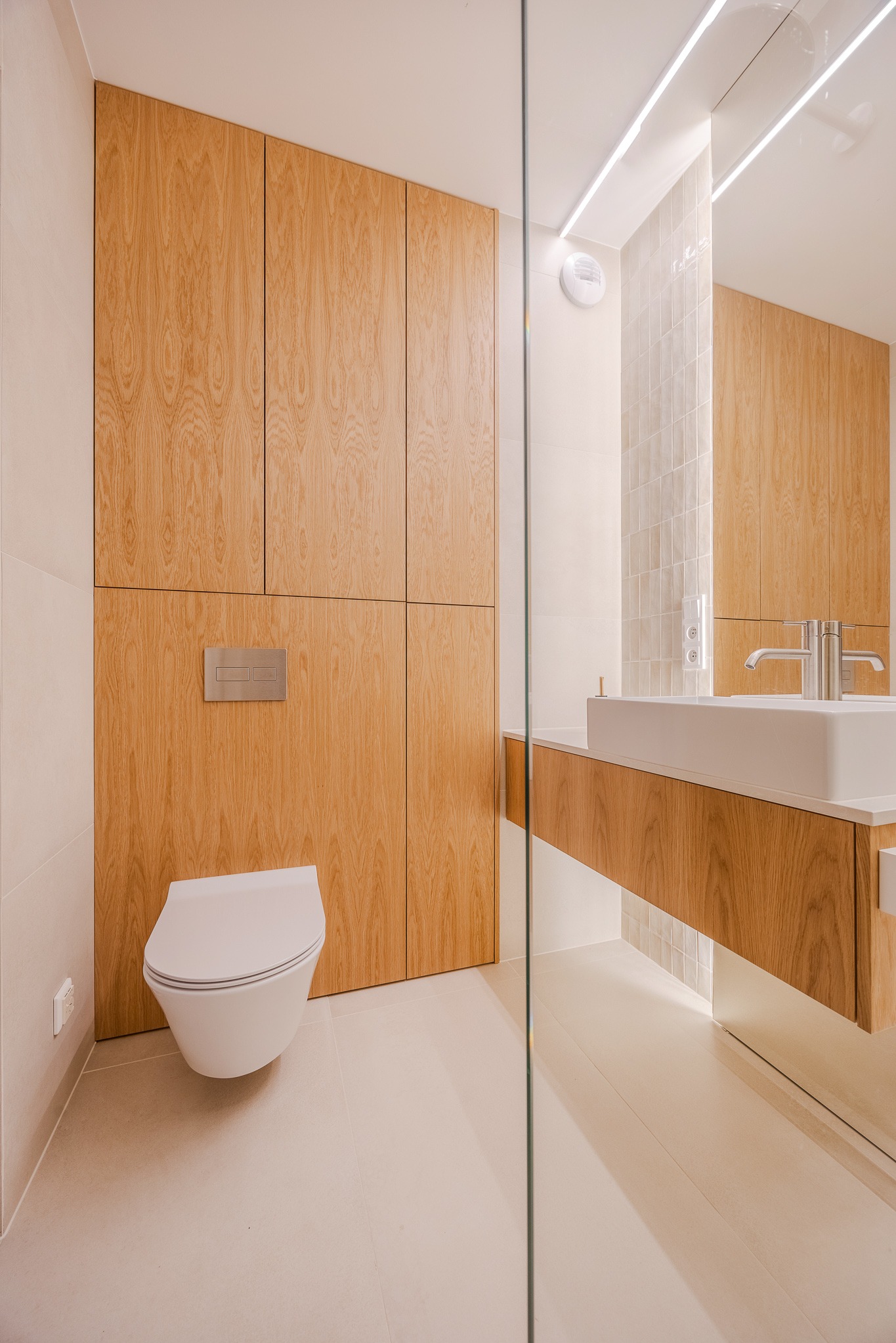 Nowoczesna łazienka z drewnianą zabudową meblową, podwieszaną toaletą i umywalką. Minimalistyczny design, jasne płytki i oświetlenie LED.