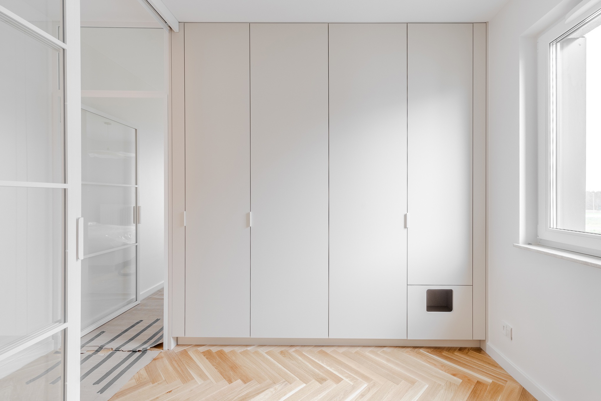 Biała szafa wnękowa z ukrytym wejściem dla kota, minimalistyczny design, parkiet w jodełkę, jasne wnętrze, okno z widokiem na zieleń.