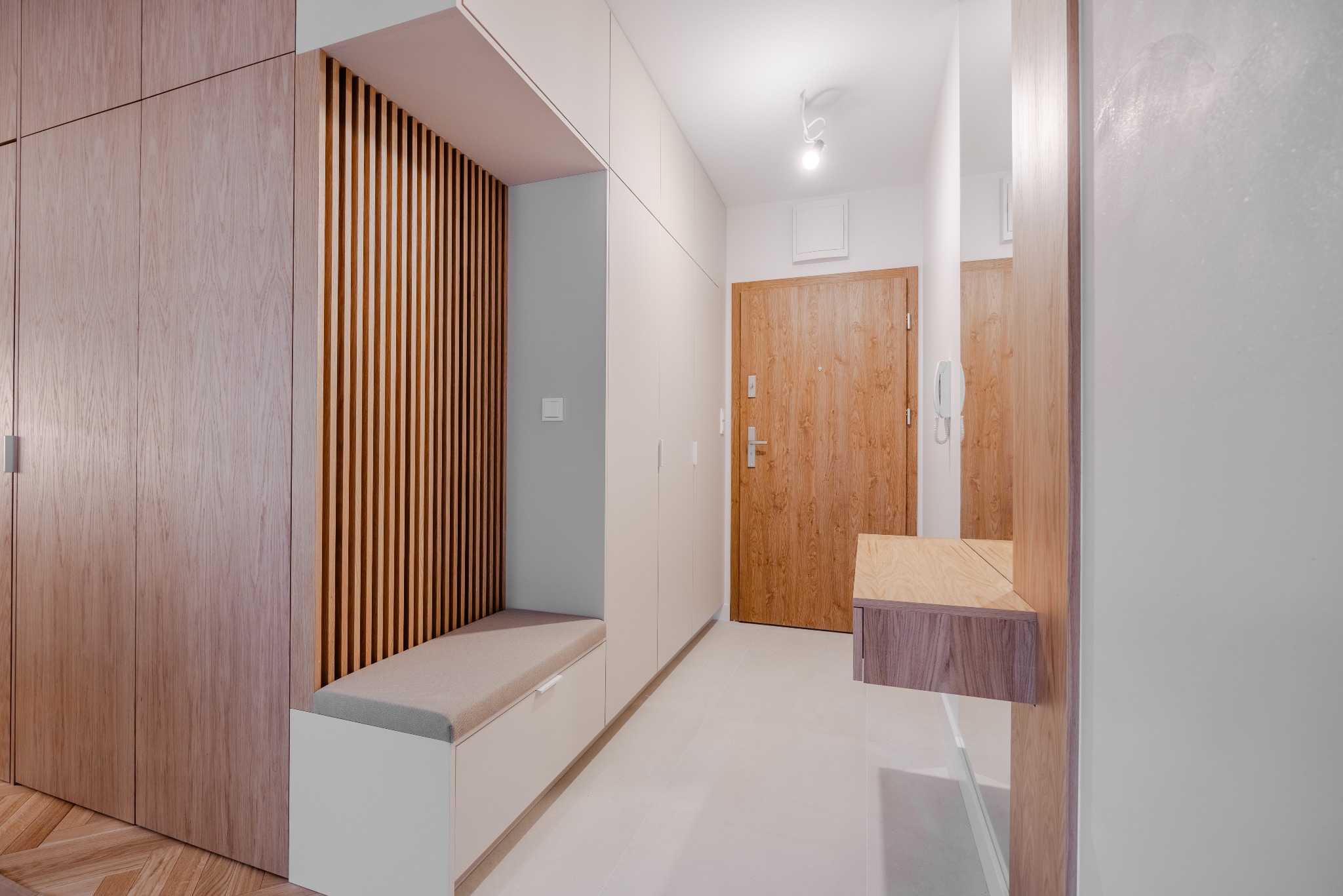 Nowoczesny przedpokój z zabudową meblową: szafy z jasnego drewna, siedzisko z szarym obiciem i dekoracyjne lamele. Minimalistyczny design, jasne wnętrze.
