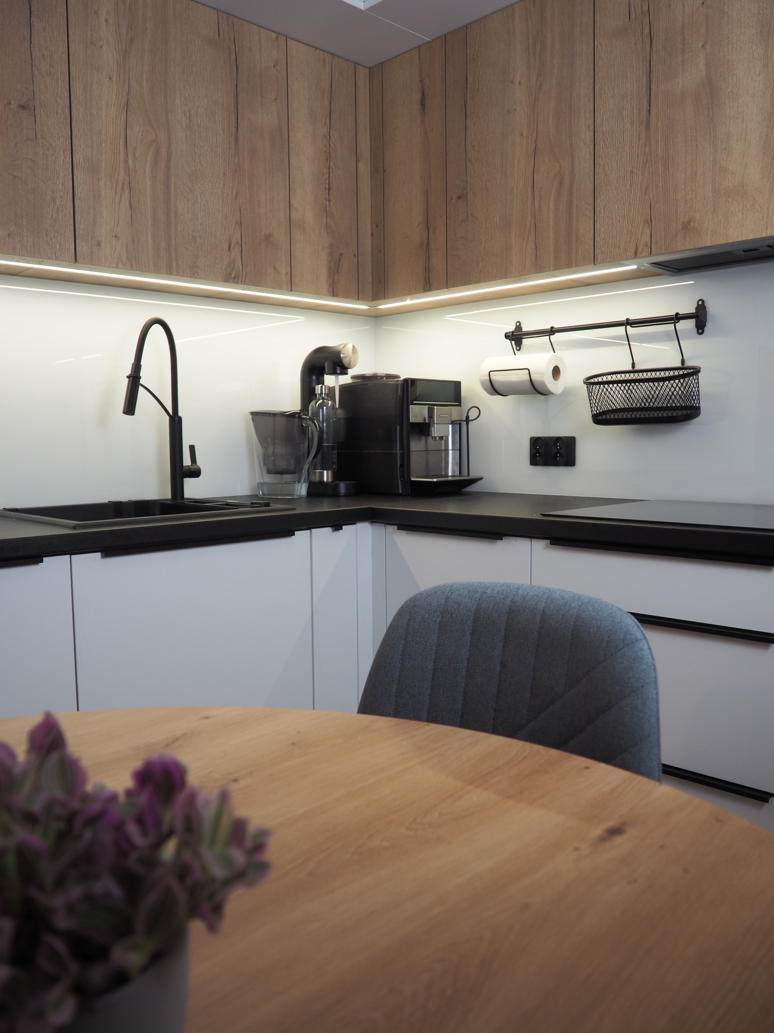 Nowoczesna kuchnia z drewnianymi szafkami górnymi i białymi dolnymi, czarnym blatem i armaturą. Na blacie ekspres do kawy i akcesoria. W tle okrągły stół z krzesłem.