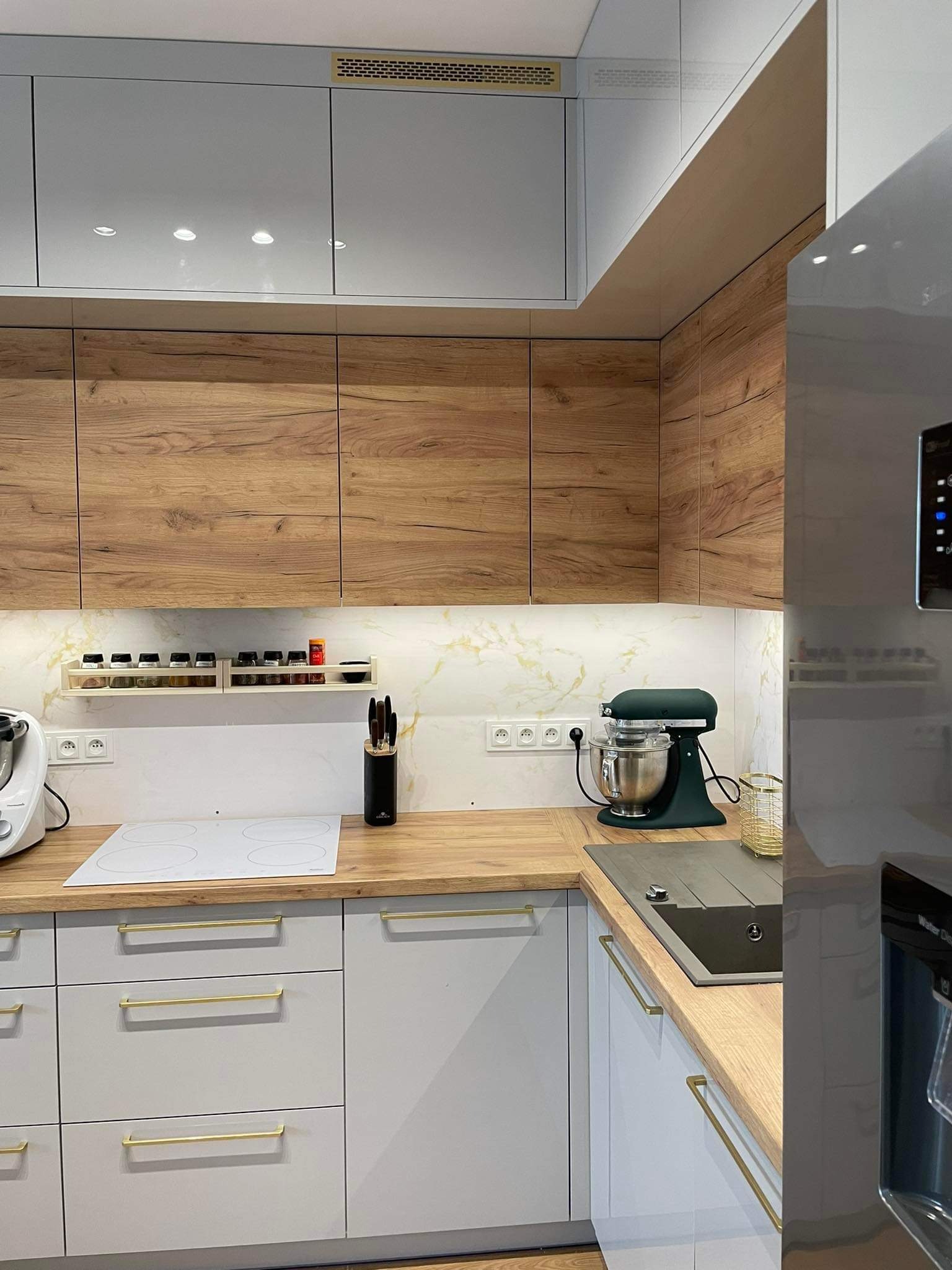 Jasnoszara kuchnia z drewnianymi akcentami: blaty i górne szafki z wyraźnym rysunkiem drewna. Złote uchwyty, biała płyta indukcyjna i marmurowy backsplash.