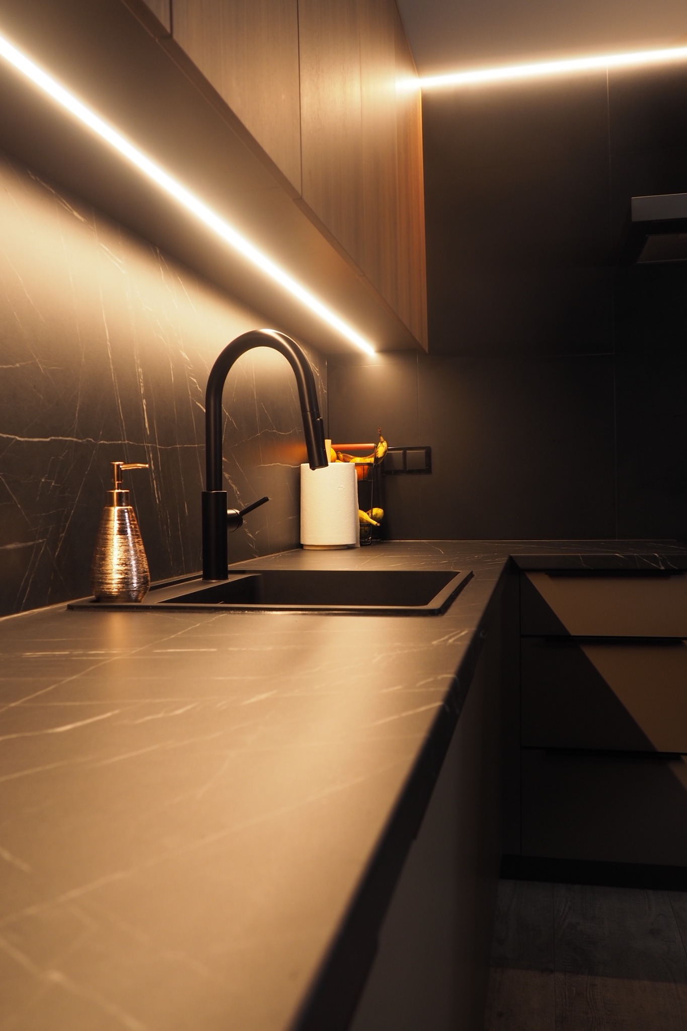 Nowoczesna, ciemna kuchnia z blatem imitującym marmur, czarną armaturą i oświetleniem LED. Dozownik na mydło w kolorze miedzianym dodaje elegancji.