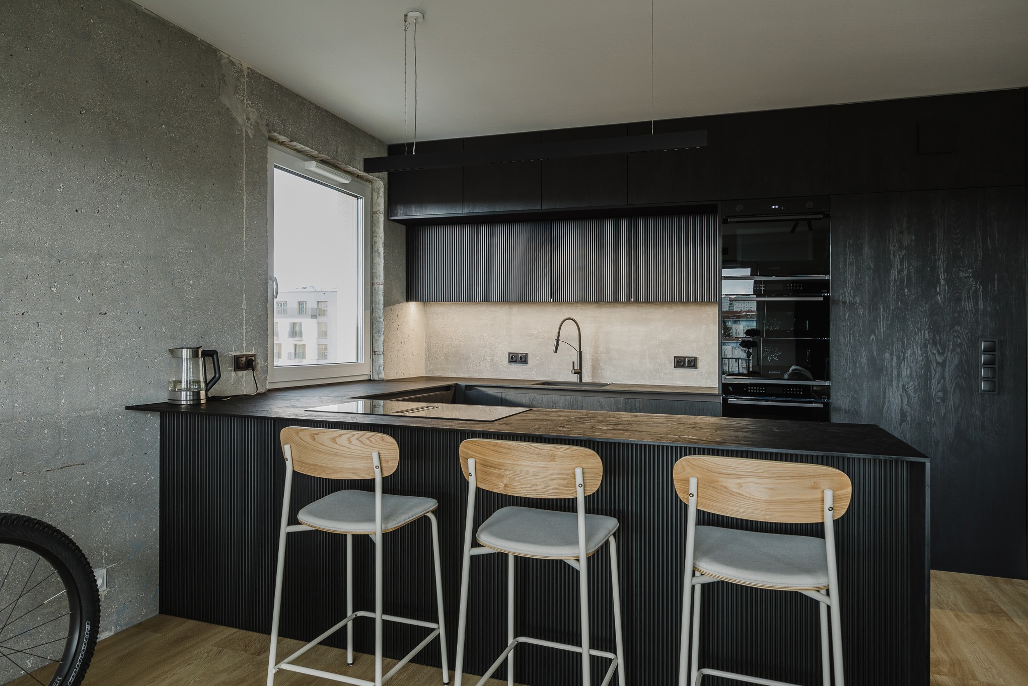 Nowoczesna, czarna kuchnia na wymiar z wyspą i trzema hokerami. Surowy beton na ścianie kontrastuje z drewnianą podłogą. Widoczne AGD w zabudowie i czajnik.