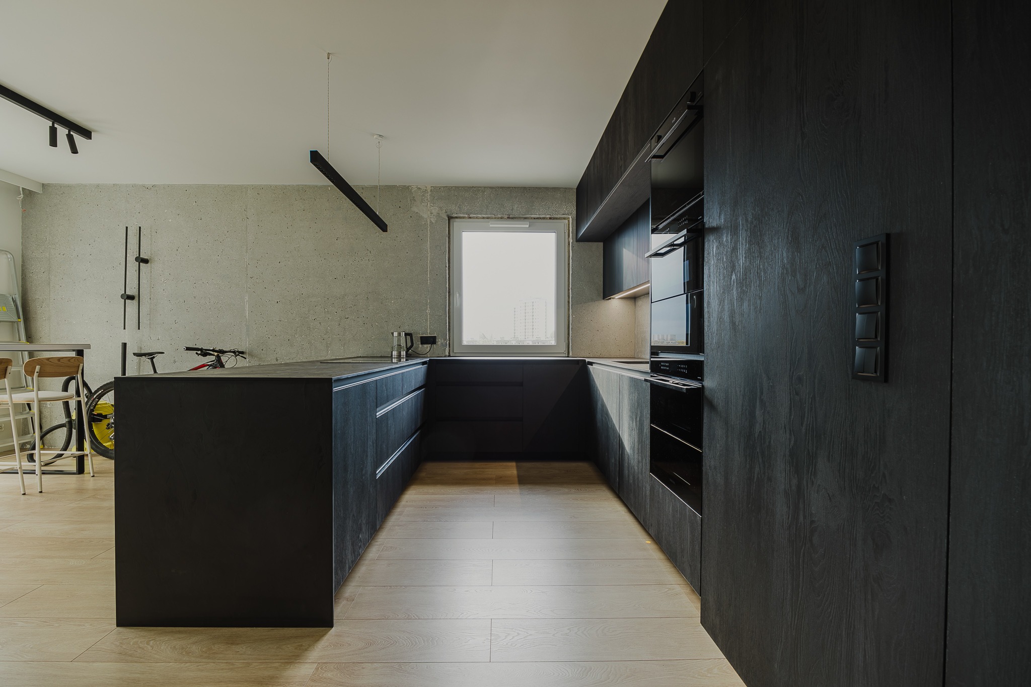 Nowoczesna, czarna kuchnia z drewnianymi frontami i betonową ścianą w tle. Widoczny rower i krzesła barowe, co dodaje wnętrzu unikalnego charakteru.