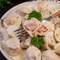 Frostcook Polska sp. z o.o - Apetyczne, gotowane pierogi z mięsem na talerzu, udekorowane zieloną pietruszką i przyprawami, jeden przekrojony widelcem, na ciemnym tle.