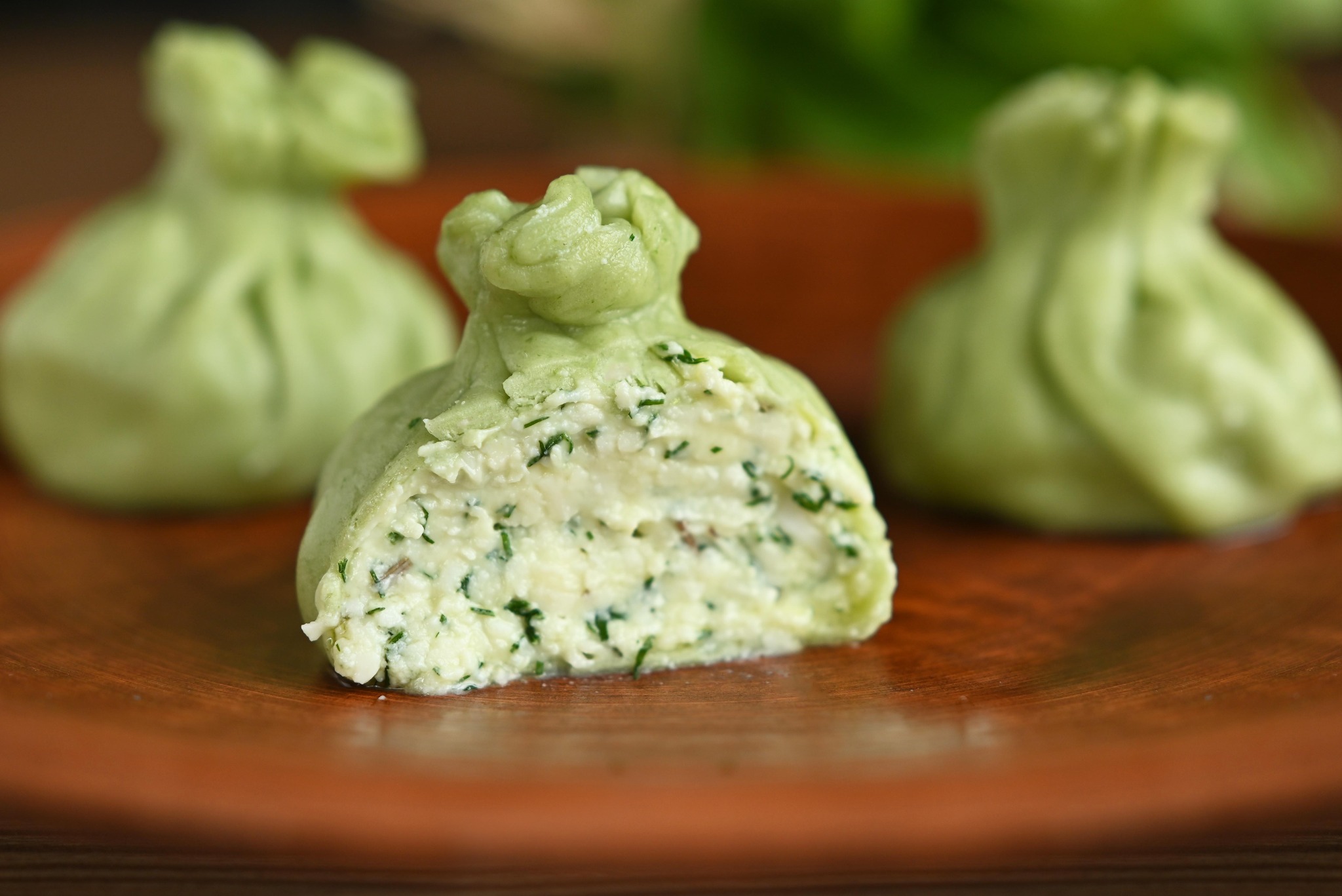 Zielone, przekrojone chinkali z farszem serowym i ziołami na brązowym talerzu, z dwoma chinkali w tle. Delikatne oświetlenie podkreśla świeżość dania.