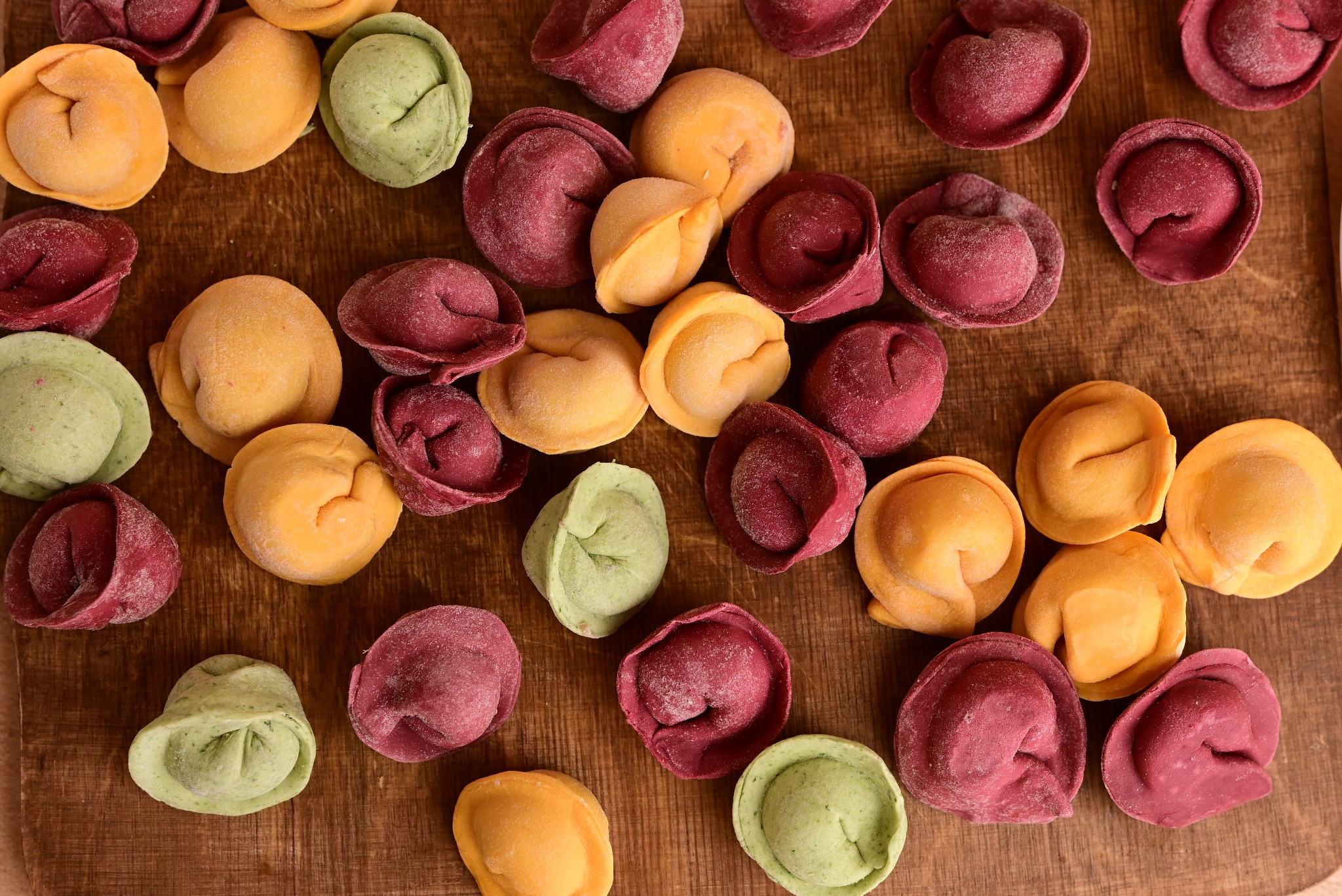 Ręcznie robione, kolorowe pierożki (żółte, zielone, czerwone) ułożone na drewnianej desce. Delikatny puder na powierzchni dodaje im rustykalnego uroku.