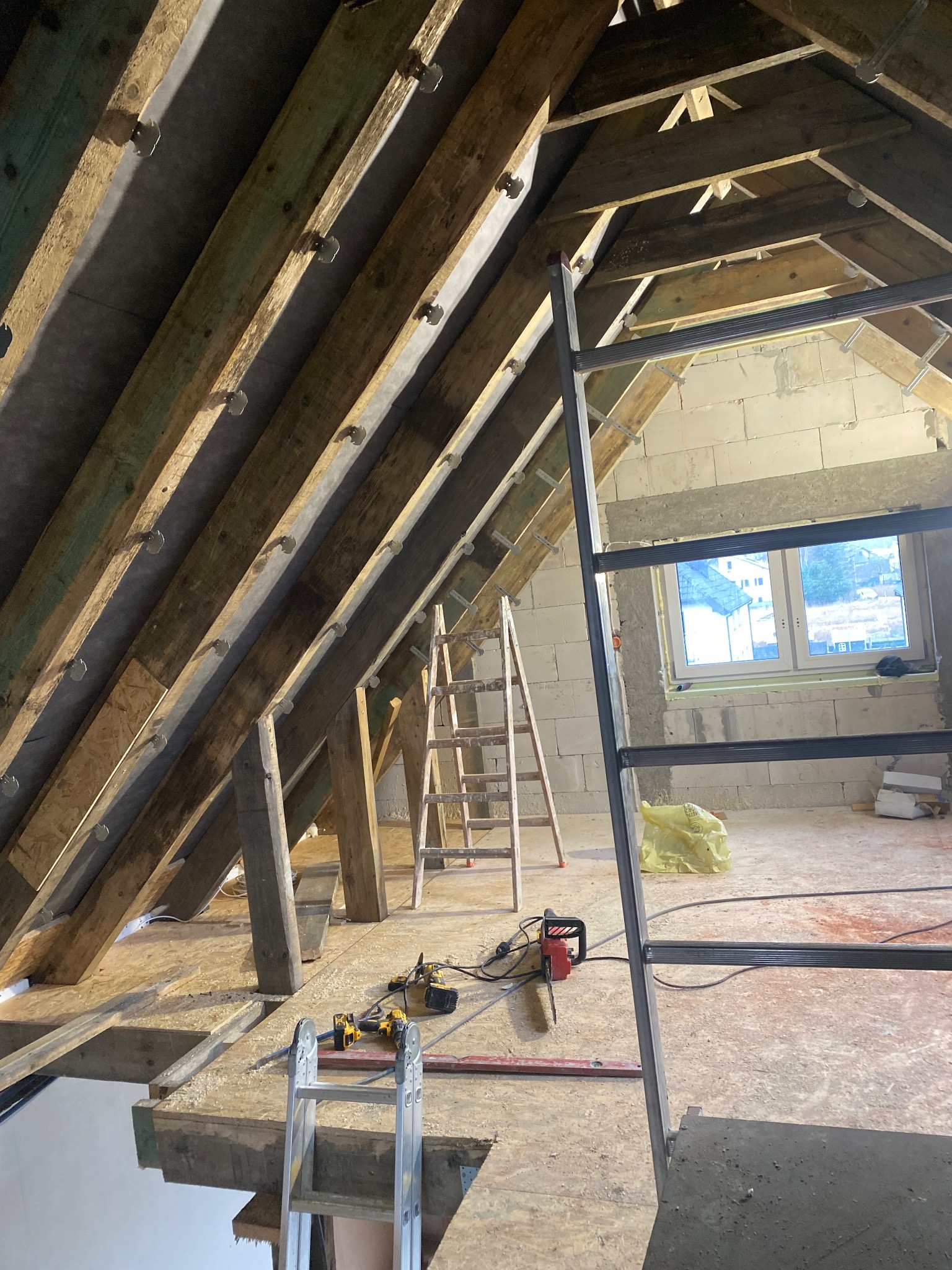 Skośny strych w trakcie adaptacji: drewniana konstrukcja dachu, okno, drabiny, narzędzia. Widok na surowy stan poddasza przed wykończeniem.