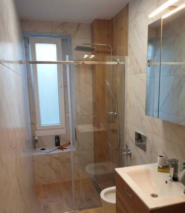 Wykończona łazienka z kabiną prysznicową typu walk-in, marmurowymi płytkami i drewnianymi akcentami. Widoczne narzędzia na parapecie okna.