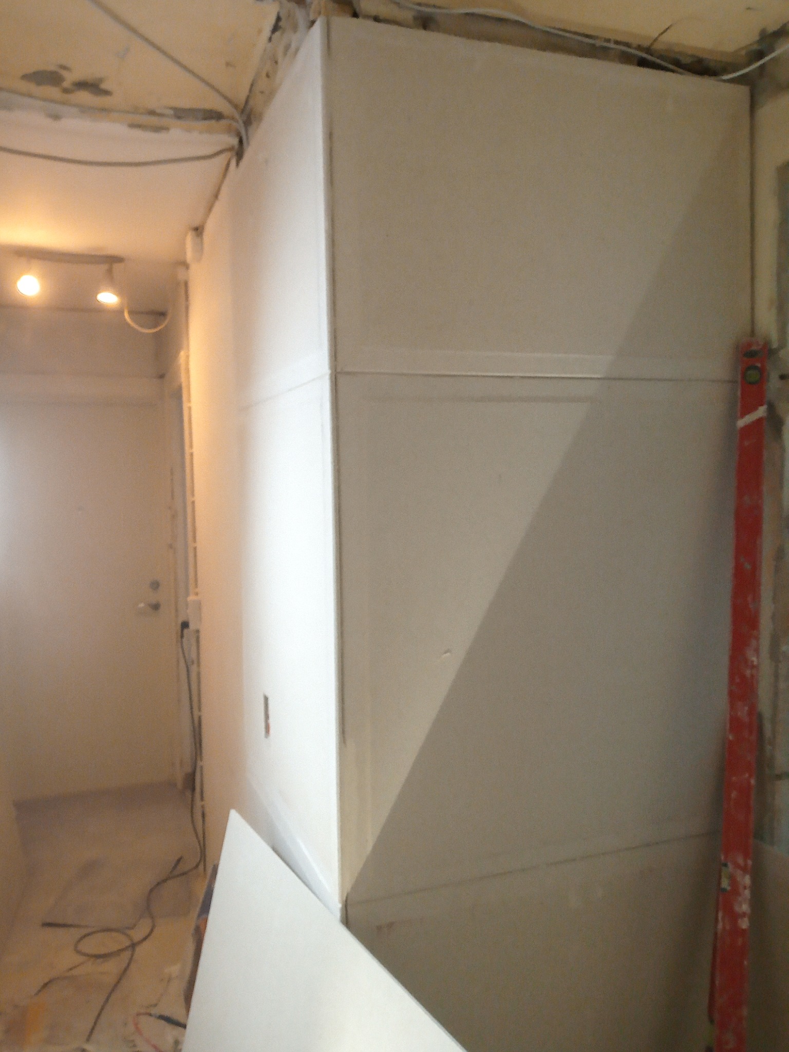 Wnętrze w trakcie prac: zabudowa z płyt kartonowo-gipsowych, widoczny poziomica, kable i fragment drzwi. Oświetlenie sufitowe.