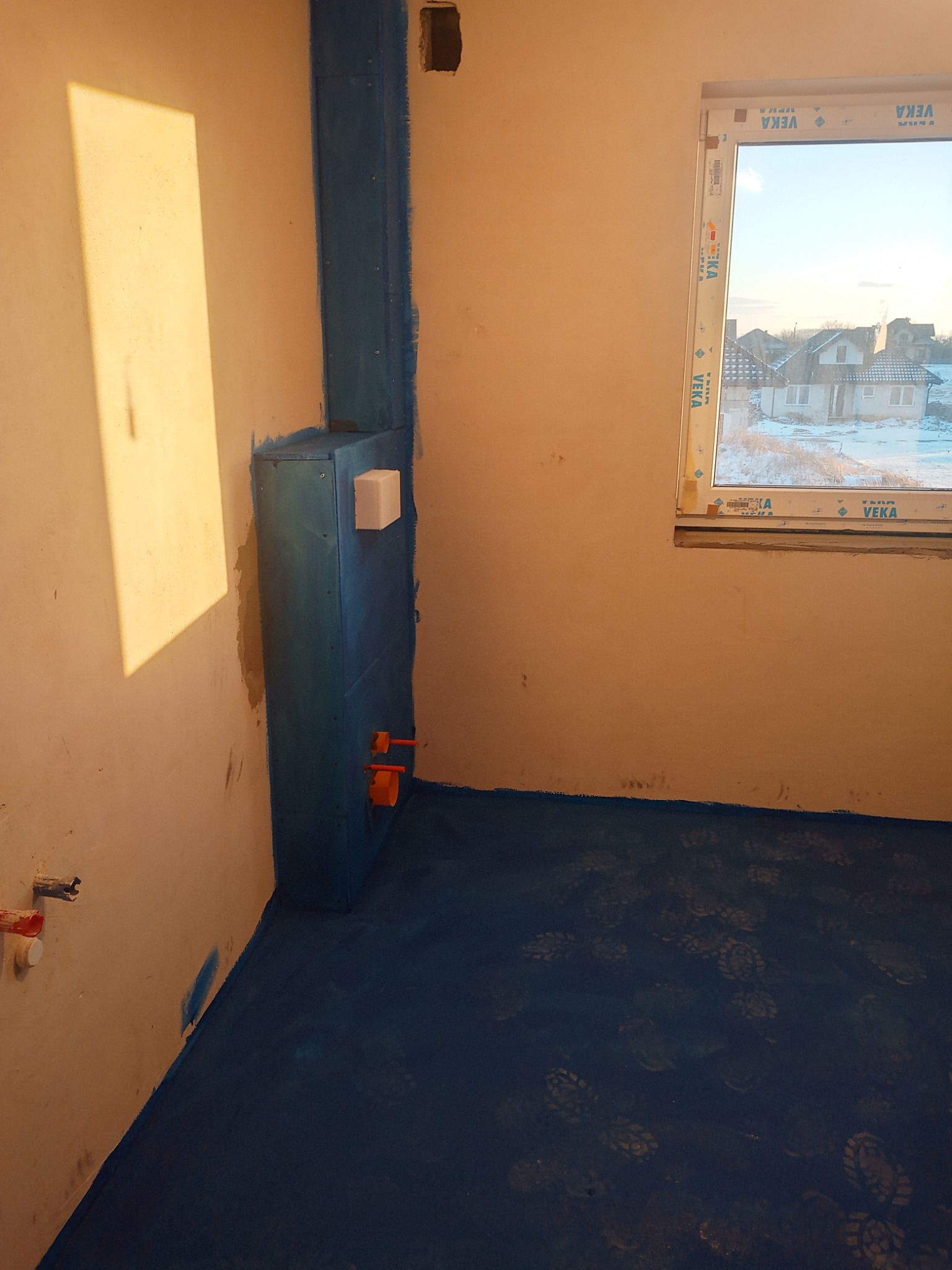 Niebieska hydroizolacja w rogu łazienki, zabezpieczająca ścianę i podłogę. Widoczne wystające rury i okno z widokiem na ośnieżone domy.