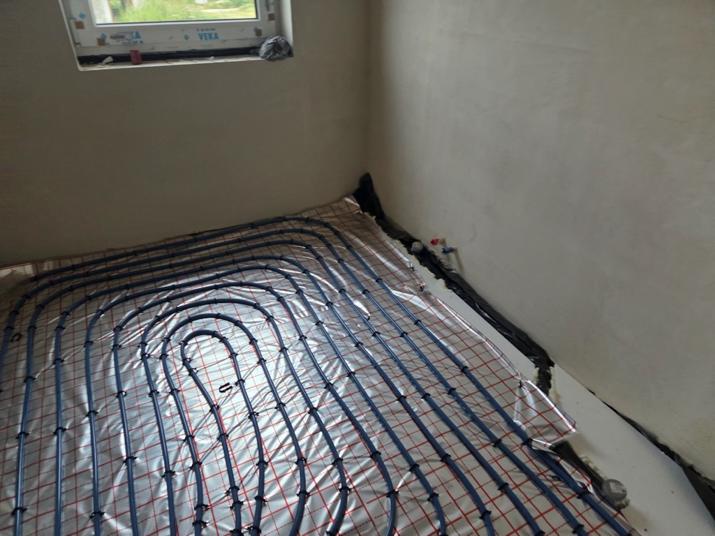 Niebieskie rury ogrzewania podłogowego ułożone na srebrnej folii z czerwoną siatką, widoczne przy ścianie z oknem, przed wylaniem posadzki.