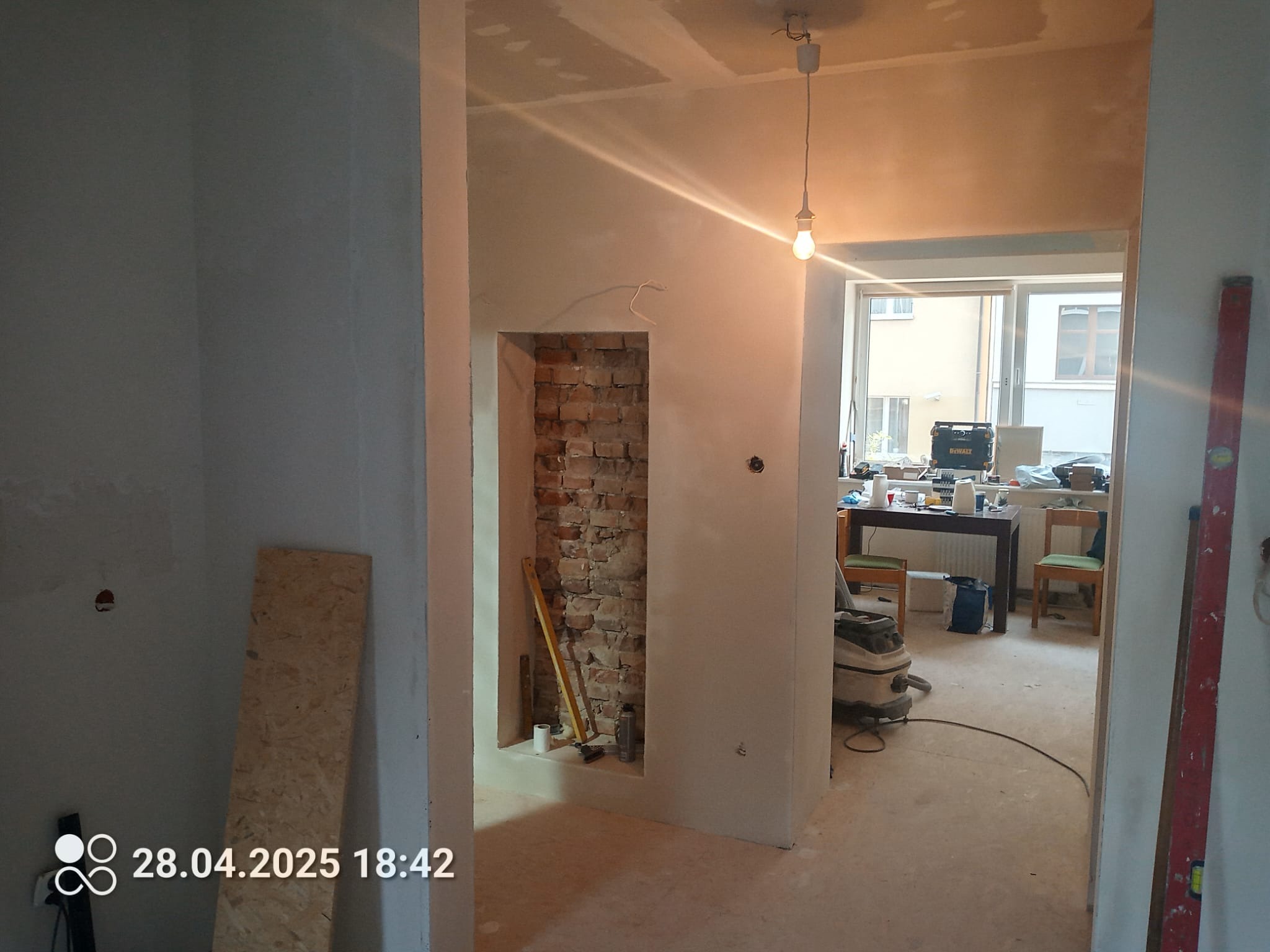 Wnętrze w trakcie remontu: surowe ściany przygotowane do gładzi, odsłonięta cegła we wnęce, widoczne narzędzia i materiały budowlane. Żarówka zwisa z sufitu.