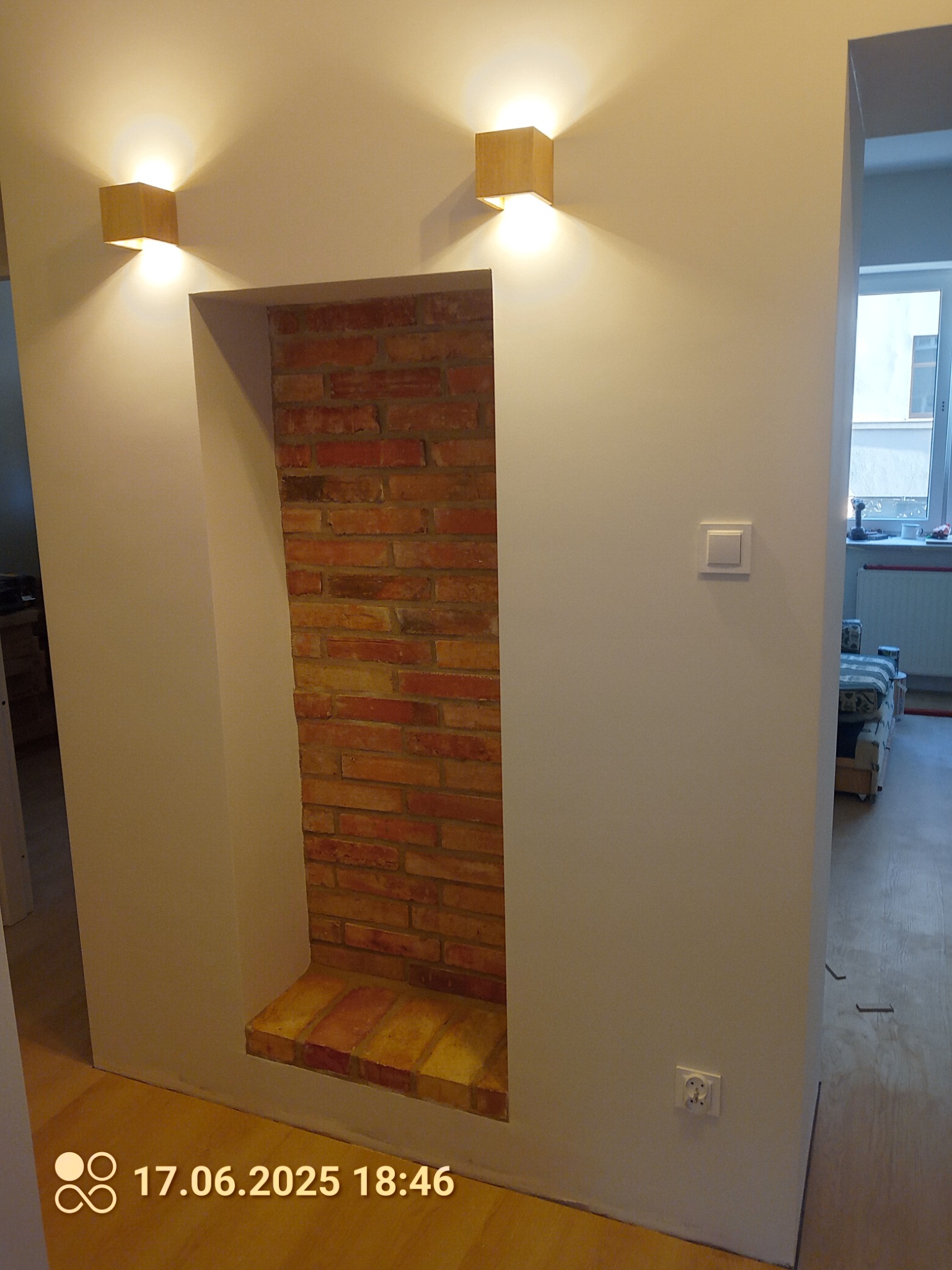 Wnęka z cegły w pomalowanej na biało ścianie, oświetlona dwoma kwadratowymi lampami. Widoczny fragment pokoju z drewnianą podłogą i gniazdko elektryczne.