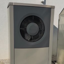 Szara pompa ciepła powietrze-woda, zainstalowana przy białej ścianie budynku, na tle trawy i kamieni. Widoczny wentylator pompy.