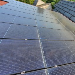 Panele fotowoltaiczne na dachu domu z widoczną konstrukcją montażową i fragmentem dachu sąsiedniego budynku w tle.