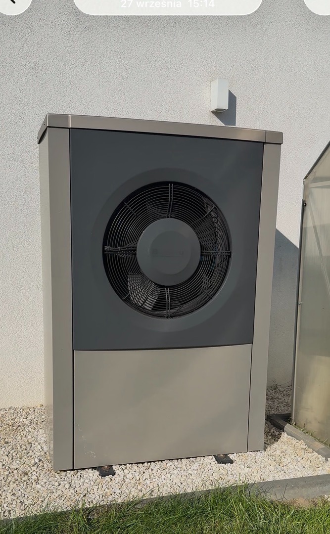 Szara pompa ciepła powietrze-woda, zainstalowana przy białej ścianie budynku, na tle trawy i kamieni. Widoczny wentylator pompy.