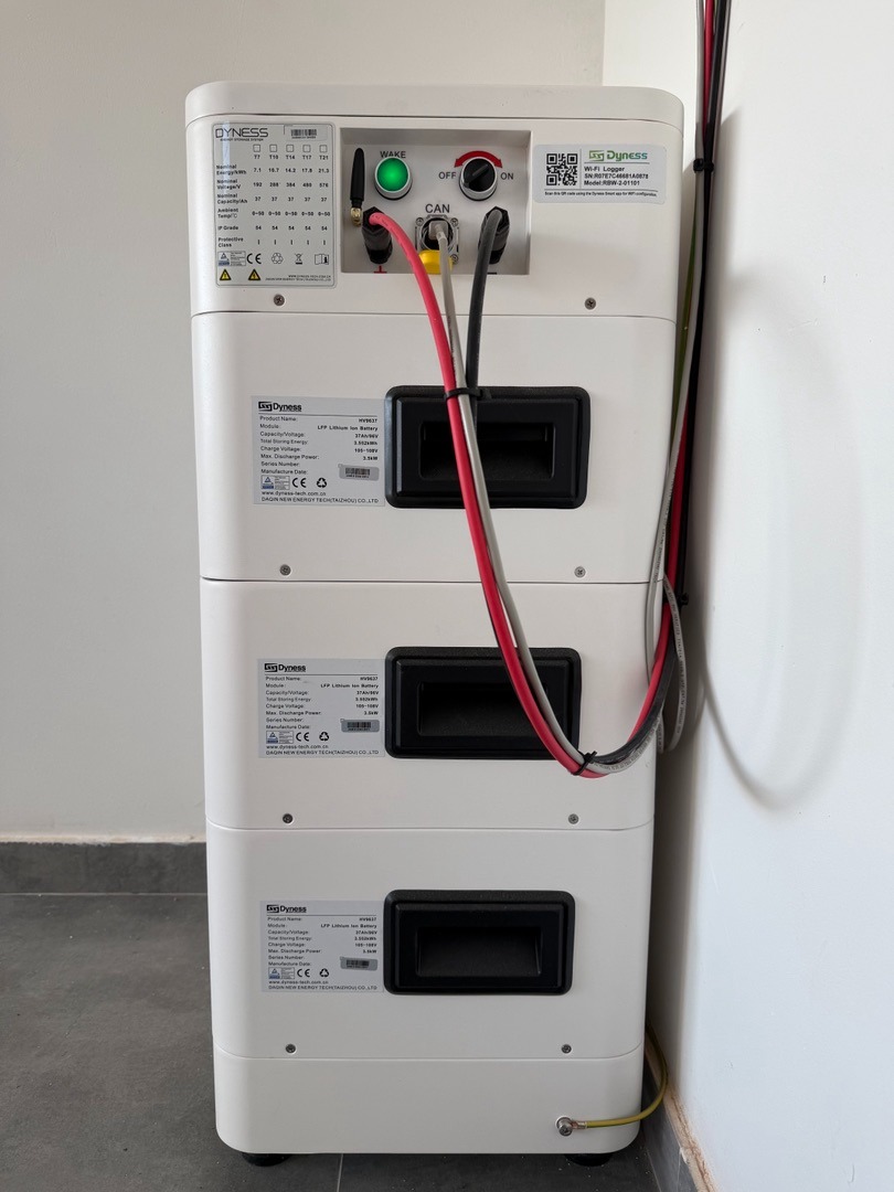 Biały, modułowy magazyn energii Dyness z widocznymi kablami i złączami, ustawiony przy białej ścianie. Widoczne etykiety i włącznik zasilania.