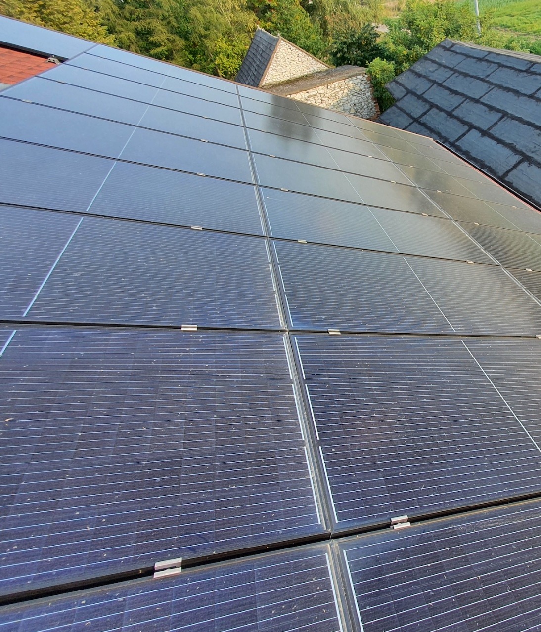 Panele fotowoltaiczne na dachu domu z widoczną konstrukcją montażową i fragmentem dachu sąsiedniego budynku w tle.