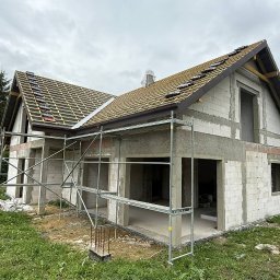 KupSobieSpokój - My budujemy. Ty żyjesz bez stresu - Surowy stan domu z białego pustaka, z częściowo ułożonym dachem z dachówki ceramicznej. Widoczne rusztowania i materiały budowlane. Etap budowy domu jednorodzinnego.