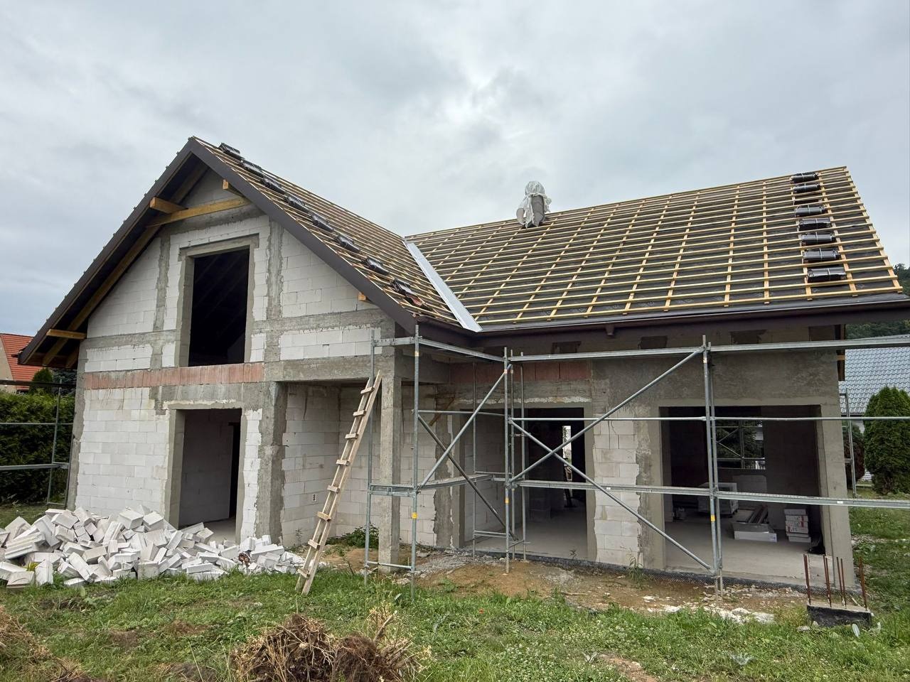 Surowy stan domu z białego bloczka, z częściowo ułożonym dachem i rusztowaniem. Na pierwszym planie sterta bloczków i drabina oparta o ścianę.