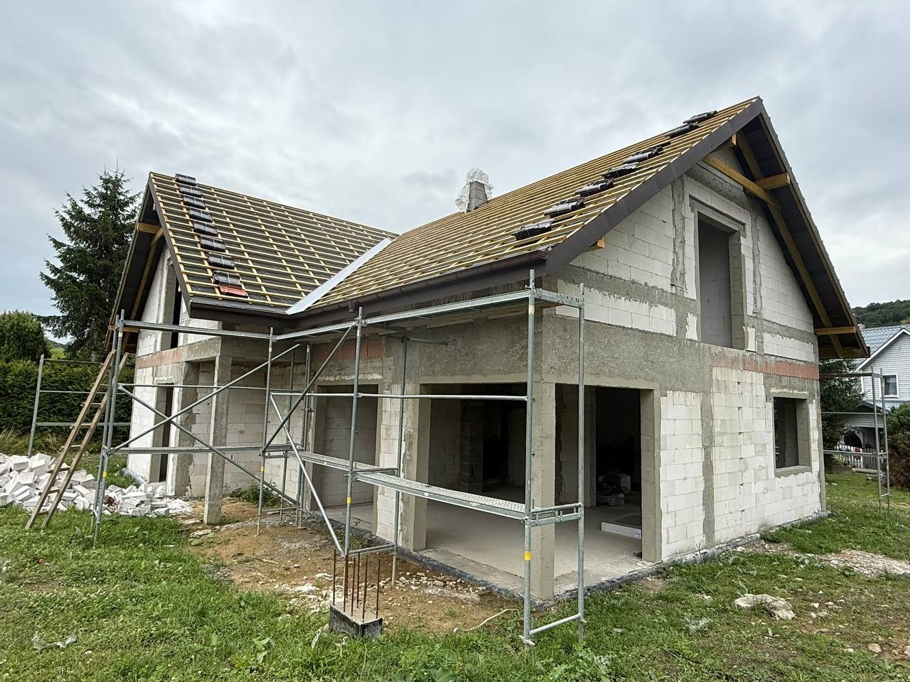 Surowy stan domu z białego pustaka, z częściowo ułożonym dachem z dachówki ceramicznej. Widoczne rusztowania i materiały budowlane. Etap budowy domu jednorodzinnego.