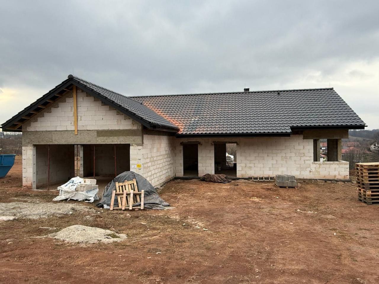 Surowy stan domu z białego pustaka z ciemną dachówką, widok z zewnątrz na plac budowy w pochmurny dzień. Częściowo otwarte przestrzenie okienne i drzwiowe.