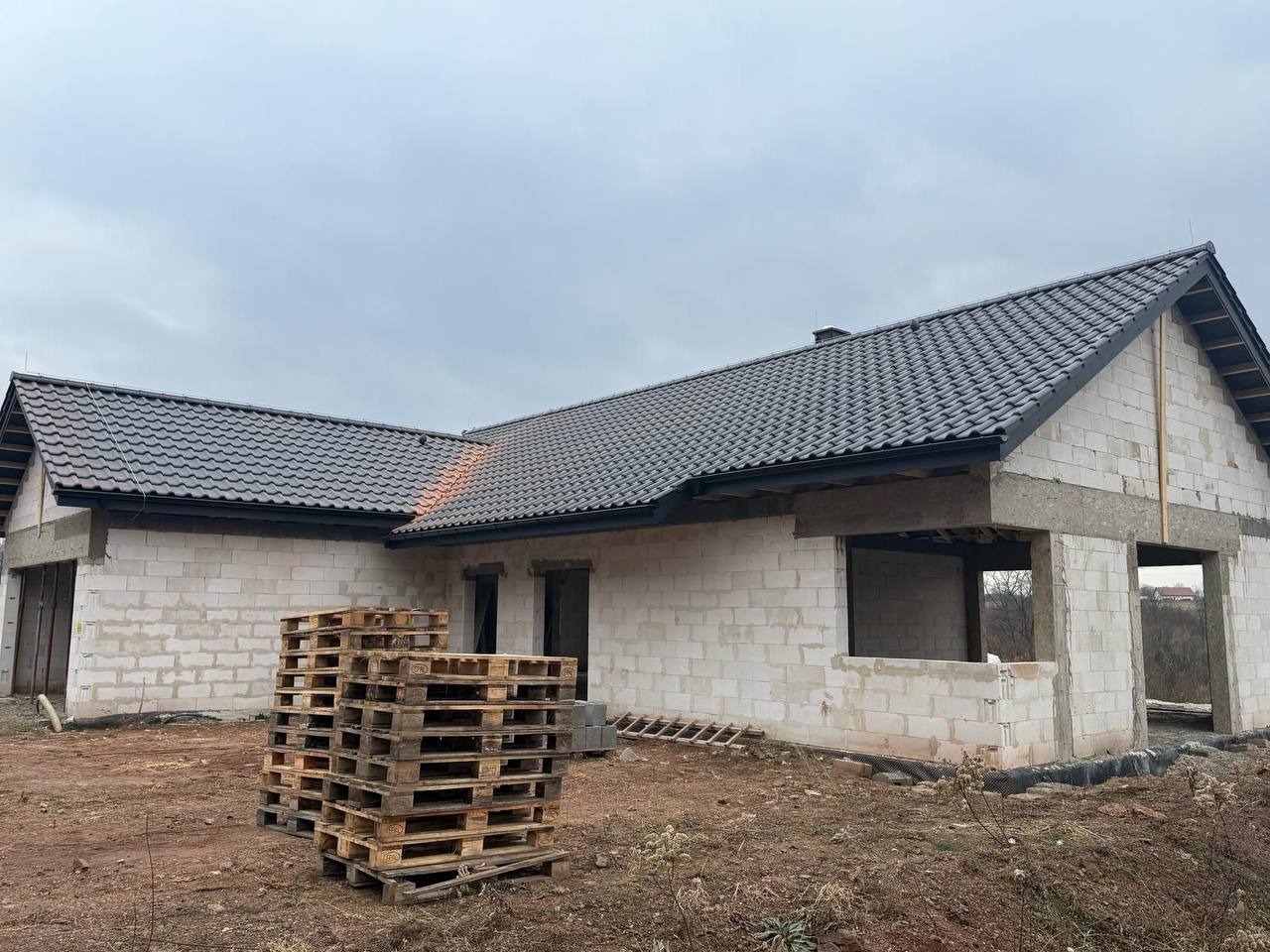 Niedokończony dom z białego pustaka z ciemnym dachem, widoczne palety na pierwszym planie, pochmurne niebo w tle. Budowa w toku, stan surowy otwarty.